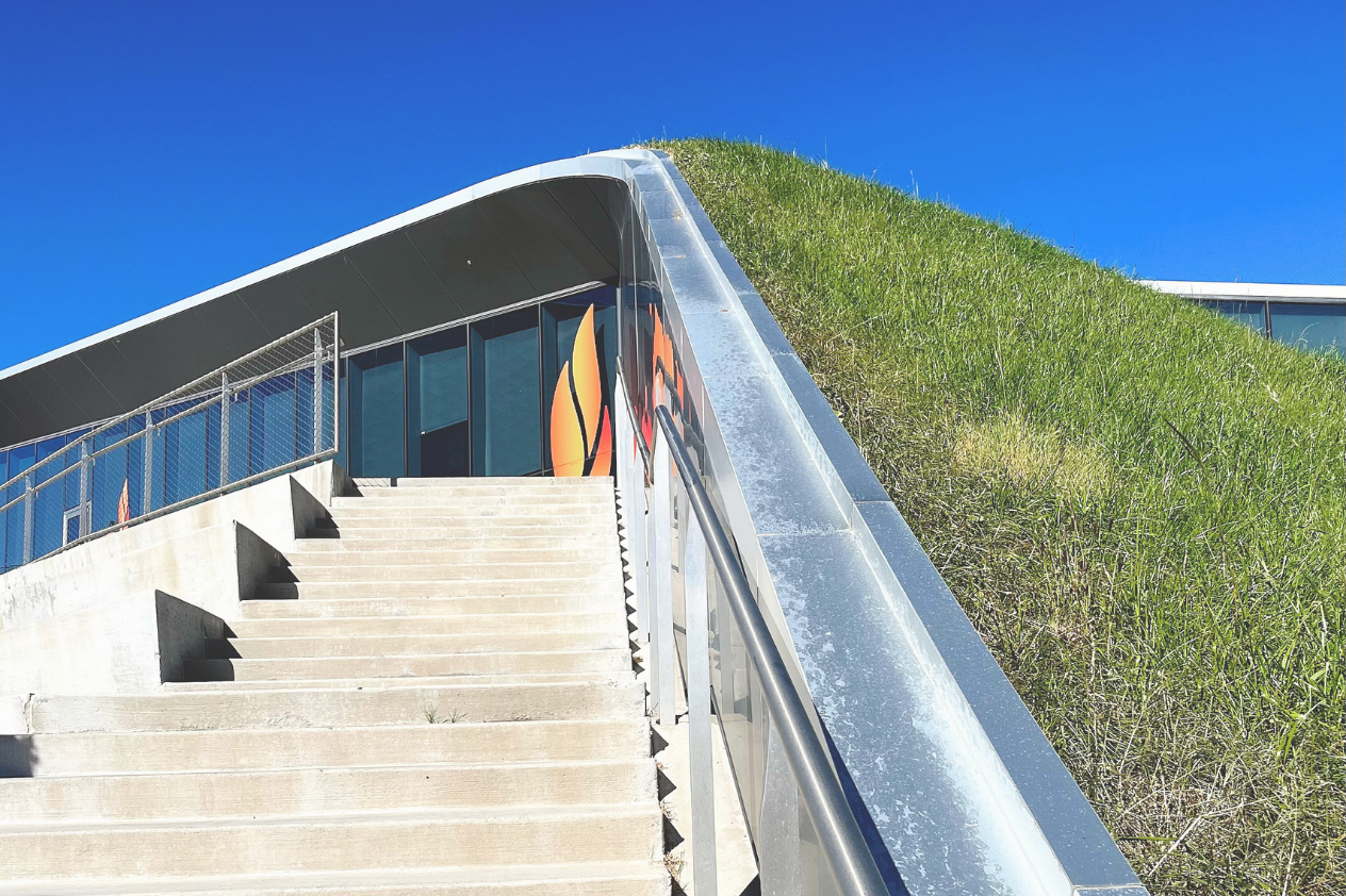 A modern building designed with a green hillside roof, featuring a curved metal edge and large glass windows. Concrete stairs lead up to the entrance, surrounded by lush grass and a clear blue sky. The design combines natural elements with contemporary architecture.