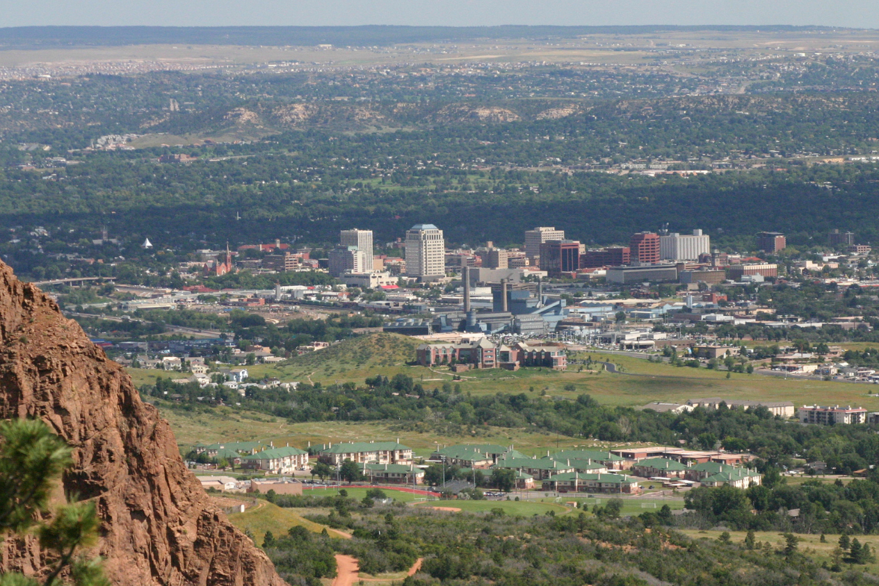 A panoramic view of a city skyline nestled in a valley, surrounded by lush green hills and mountains. The city features a mix of modern and traditional buildings, with a clear blue sky overhead and distant landscapes visible in the background.