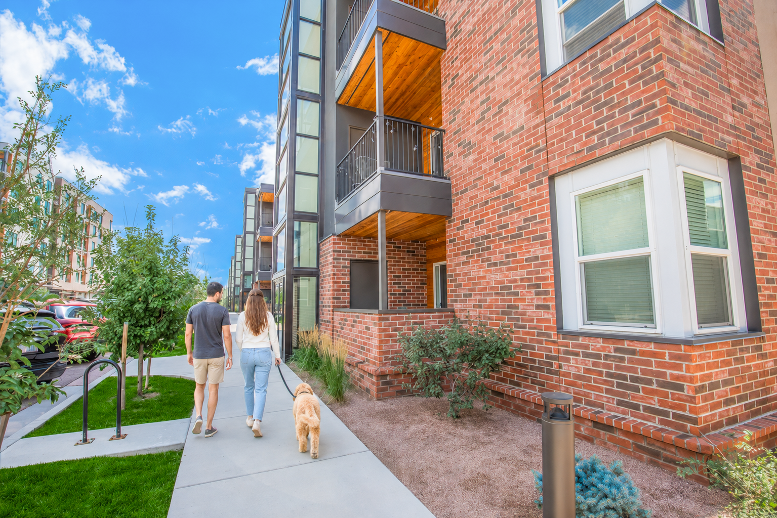A couple walks a dog along a sidewalk beside modern brick apartment buildings. The scene features green landscaping, blue skies, and a bright atmosphere, creating a welcoming residential environment.