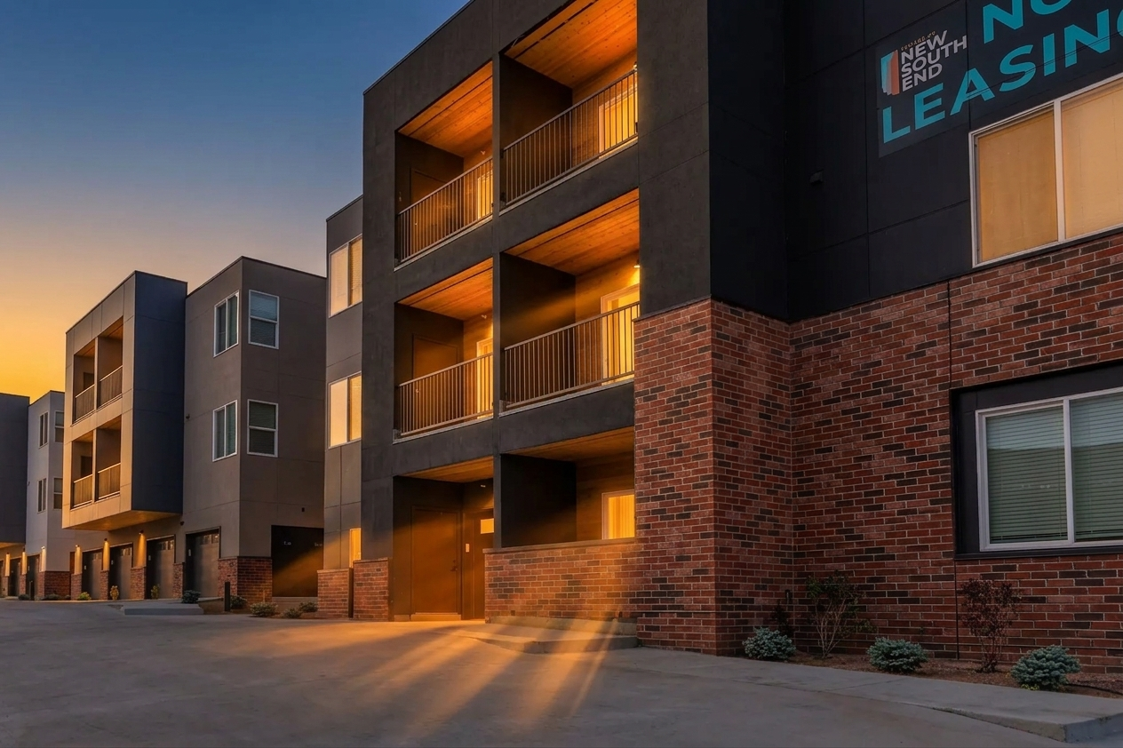 Modern apartment building with a three-story structure featuring balconies. The exterior combines dark materials with brick accents and has large windows. Soft evening light illuminates the scene, creating a warm atmosphere. A sign on the building indicates availability for leasing.
