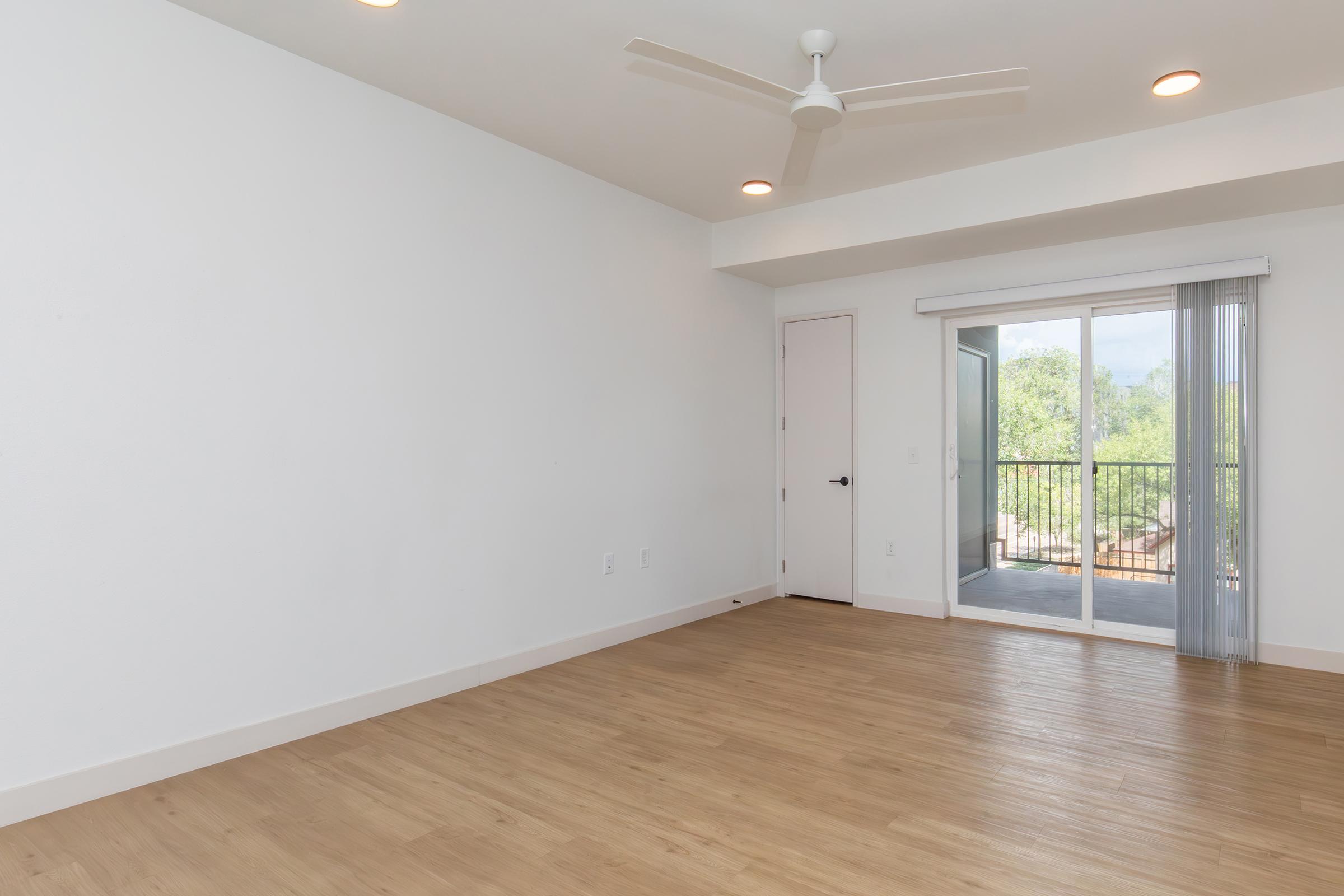 A spacious, empty room with light wooden flooring, white walls, and a ceiling fan. There is a sliding glass door leading to a balcony with a view of greenery. Natural light fills the space, creating a bright, airy atmosphere.