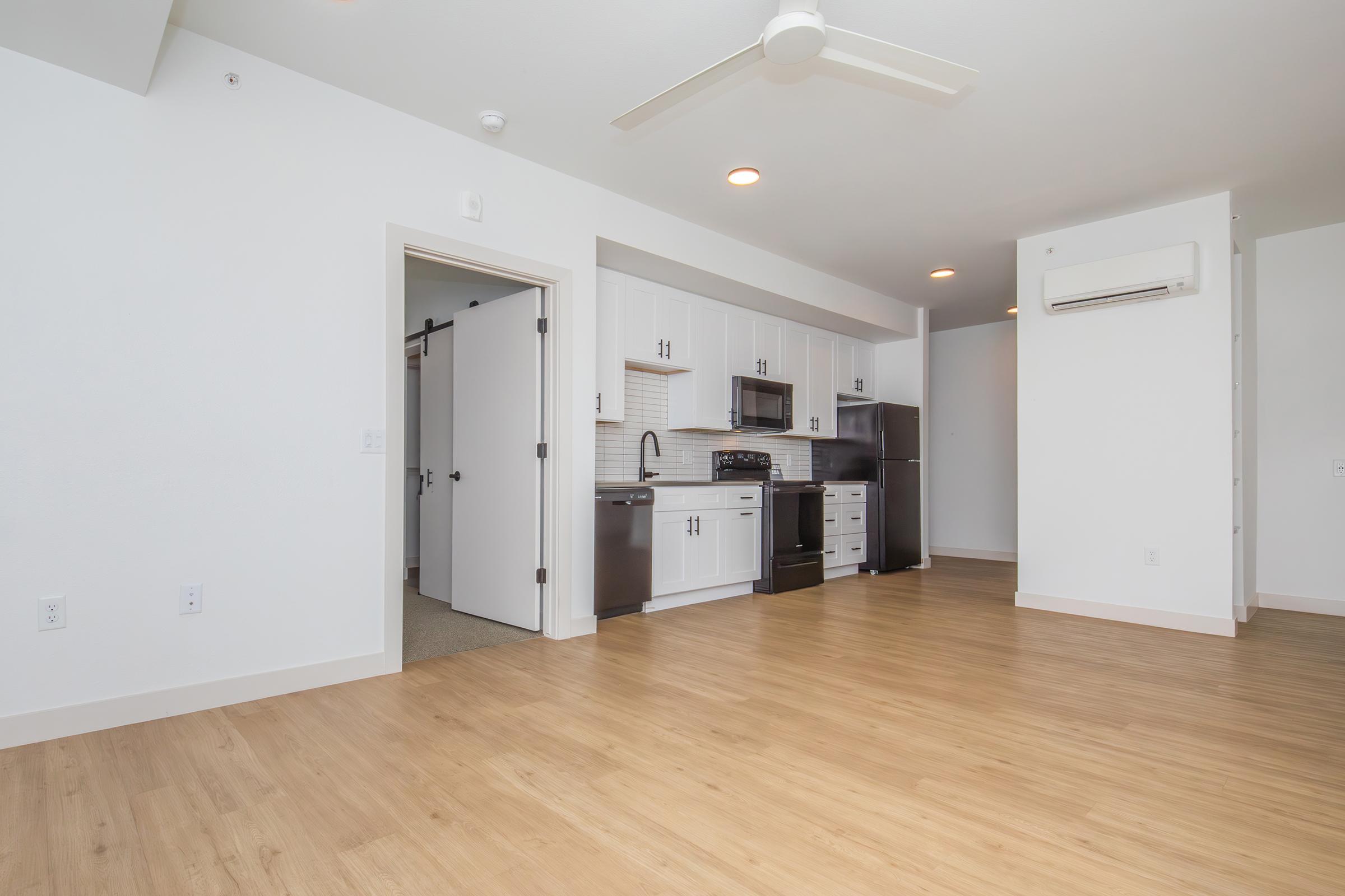A modern, open-concept apartment interior featuring light wood flooring. The kitchen is equipped with stainless steel appliances, white cabinetry, and a tiled backsplash. There is a large window letting in natural light, and a ceiling fan visible. The space is uncluttered, emphasizing a minimalist design.