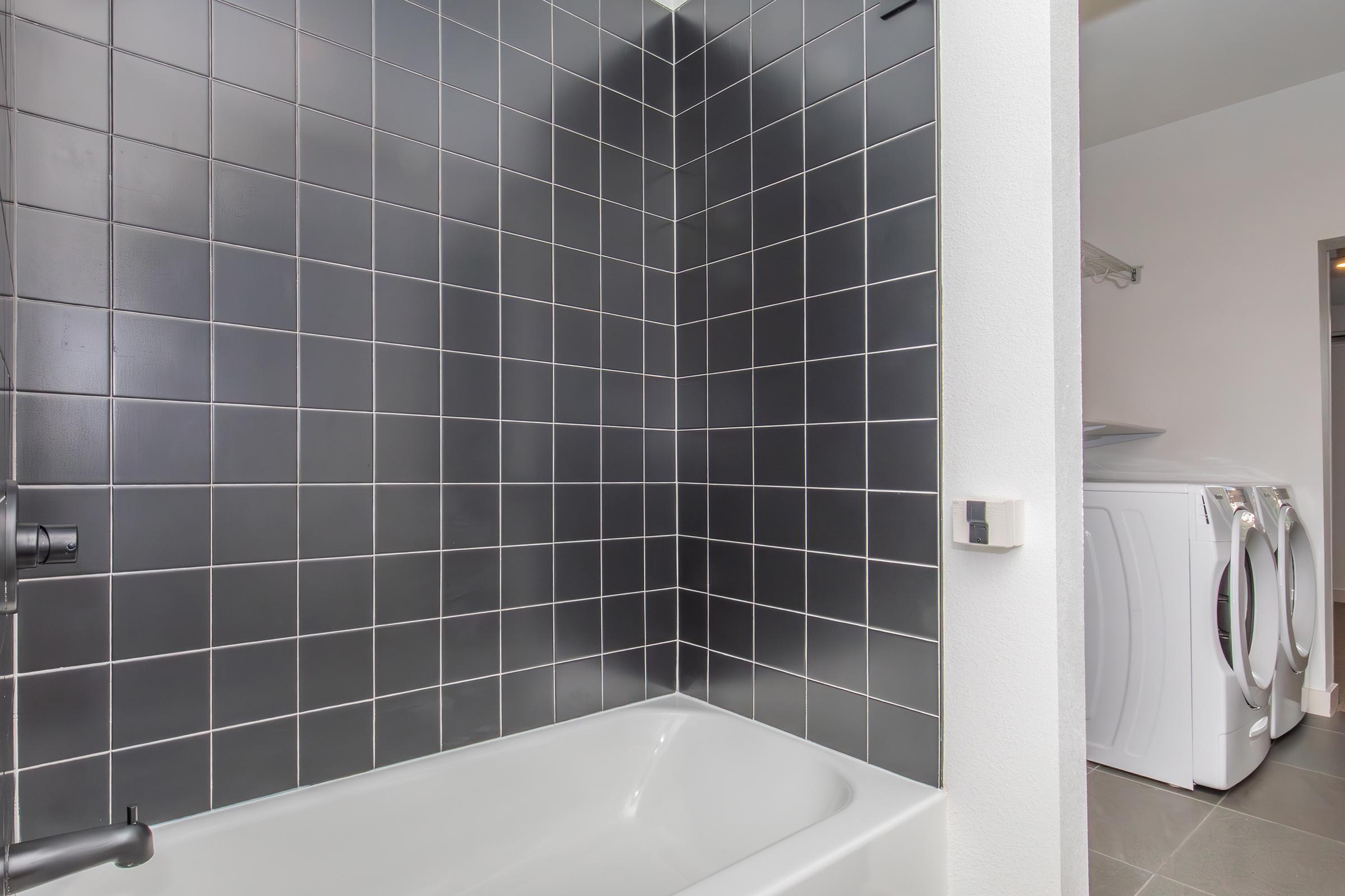 A modern bathroom featuring a sleek black tiled shower wall, a white bathtub, and a laundry area with stacked white appliances visible in the background. The overall aesthetic is clean and contemporary, with a minimalistic design.