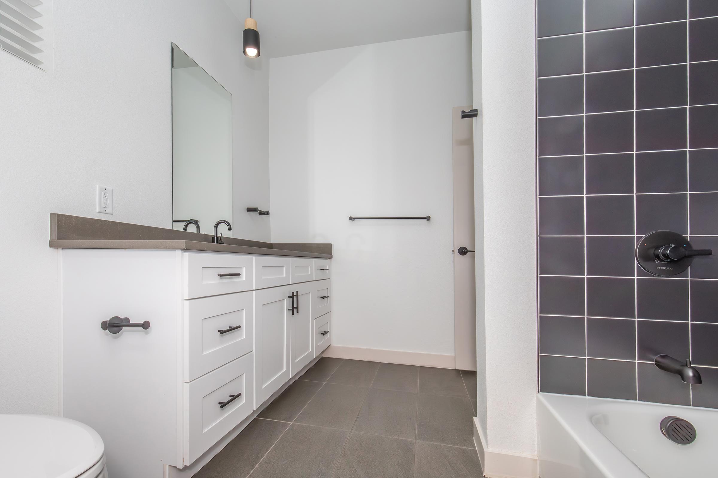 A modern bathroom featuring a white vanity with multiple drawers and dual sinks, a large mirror, and a sleek black tiled shower area. The walls are painted white, and the floor is tiled in a neutral gray. There is also a toilet visible in the corner, providing a clean and contemporary aesthetic.