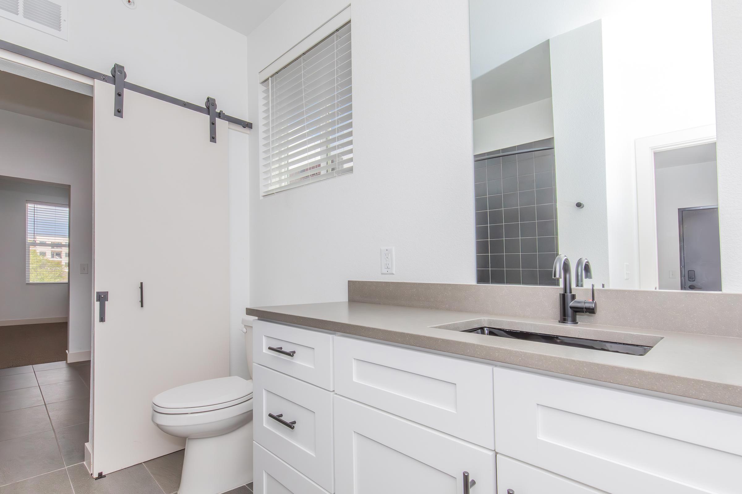 Modern bathroom featuring a white vanity with a sink, brushed nickel faucet, and large mirror. There is a sliding barn door partially open leading to a room, gray tiled shower area, and a tasteful color scheme with natural light coming from a window. The flooring is light-colored tiles.