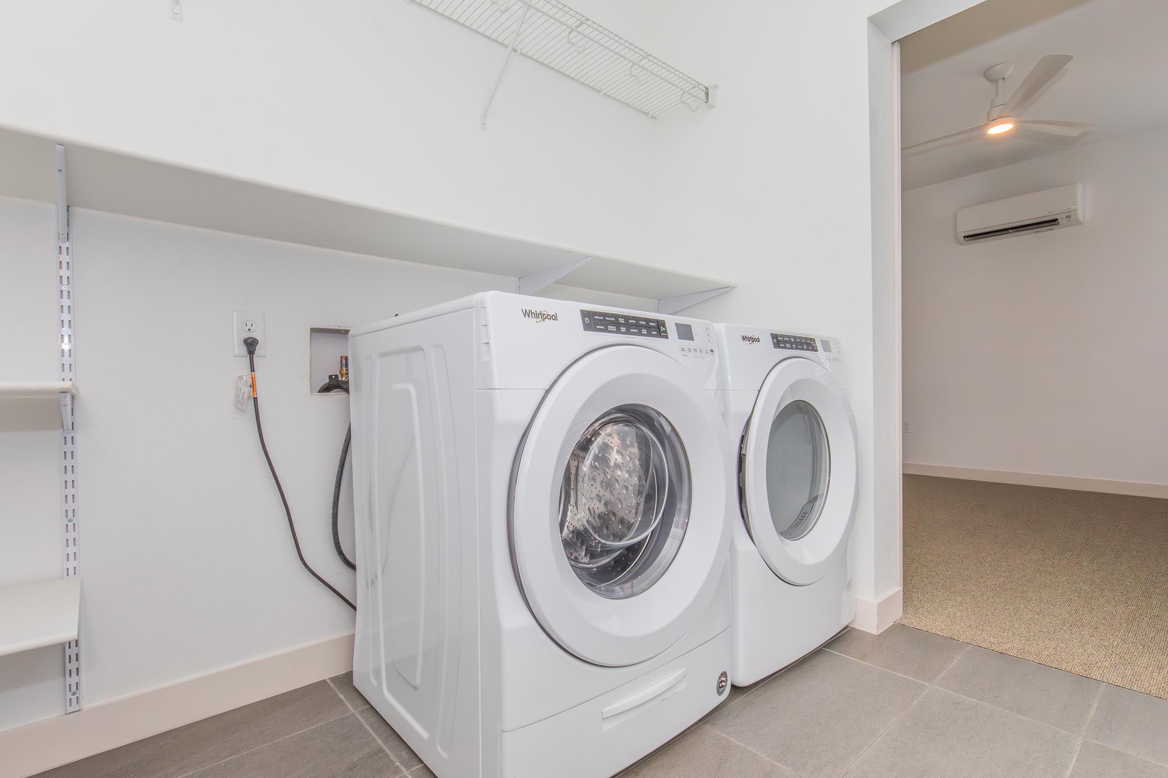 A bright laundry room featuring a pair of white front-loading washing machines side by side. There is a shelf above for storage and a ceiling fan above for ventilation. The floor is tiled with a light gray color, and the walls are painted white, creating a clean and modern aesthetic.