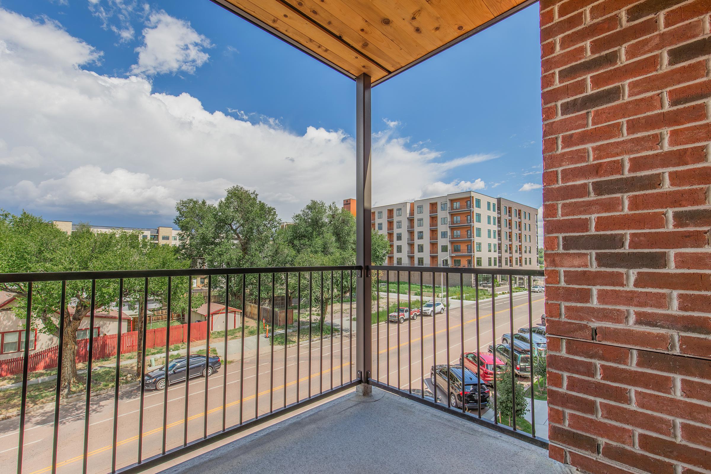 View from a balcony showing a partial streetscape with parked cars, greenery, and neighboring buildings. The sky is partly cloudy, featuring blue and white clouds. The balcony railing is black, and the terrace is made of concrete.