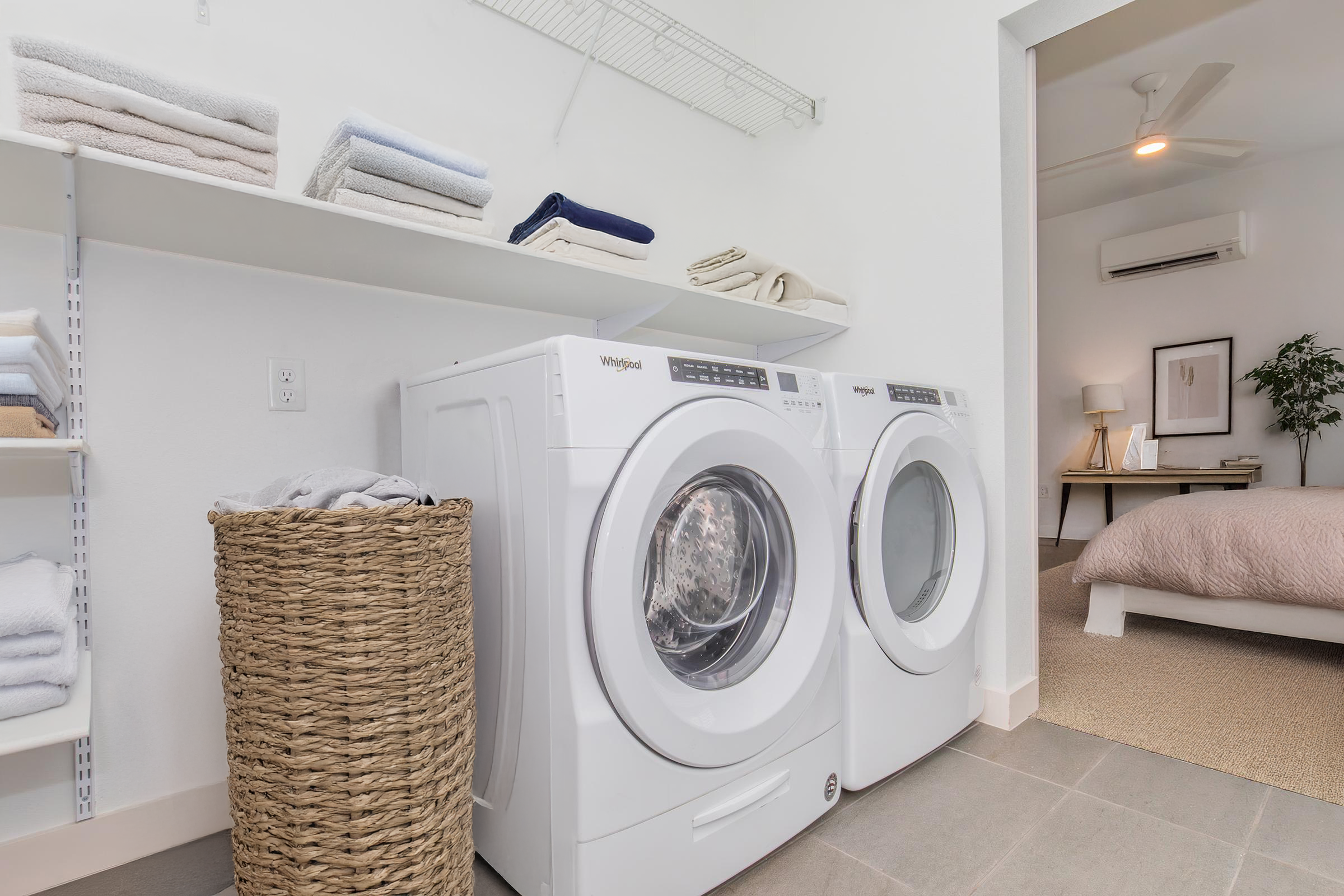 A modern laundry room featuring a pair of white front-loading washing machines with a stacked shelf above holding neatly folded towels. To the side, a woven basket filled with laundry sits on the floor, and a cozy bedroom with a light bedspread and plant is visible in the background.