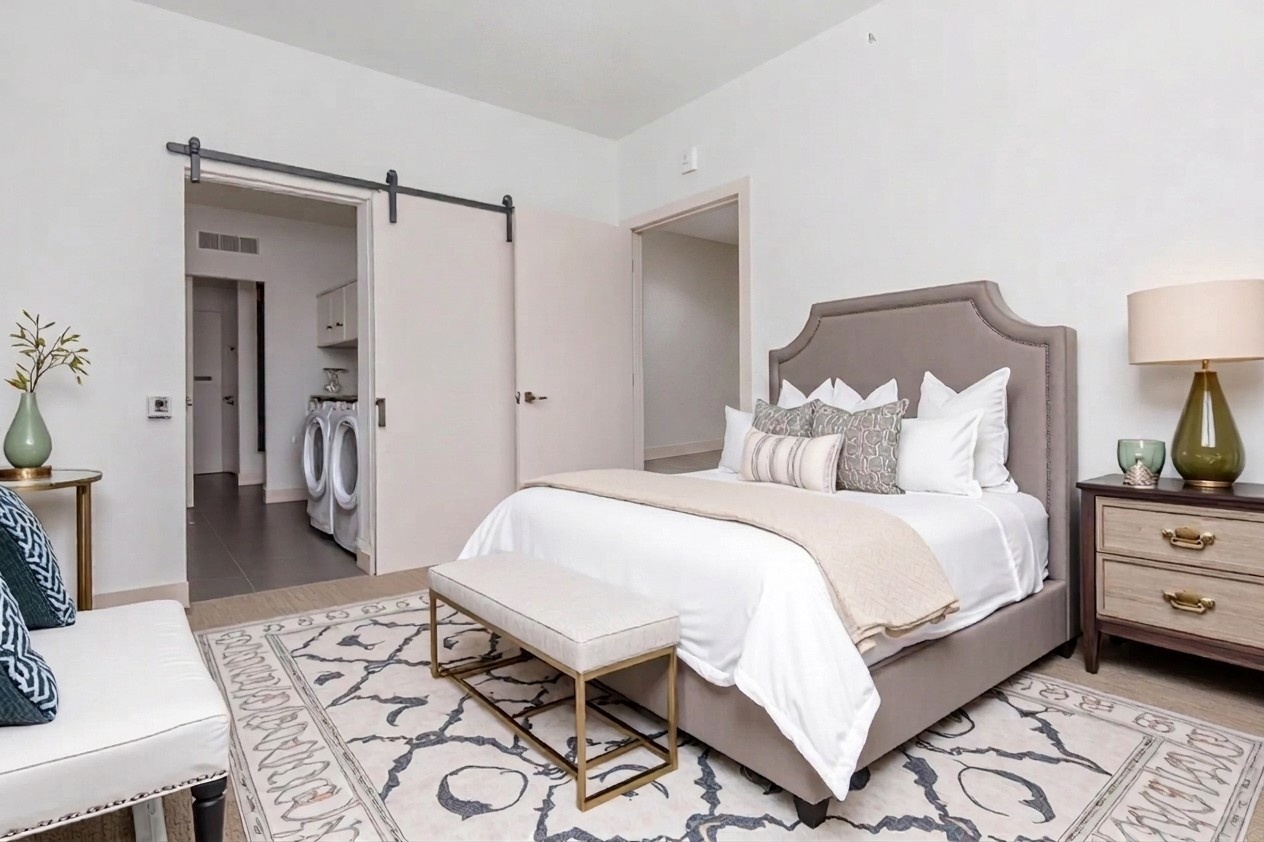 A stylish bedroom featuring a plush bed with decorative pillows, a beige throw blanket, and a bench at the foot. The room includes a nightstand with a lamp, a patterned area rug, and a cozy armchair. In the background, there's a doorway leading to a laundry area. The walls are painted in soft, neutral tones.