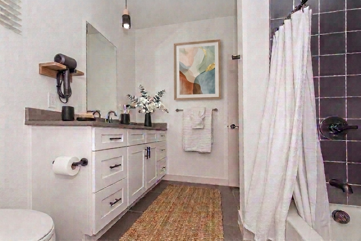 A modern bathroom featuring a white vanity with multiple drawers, a large mirror, and decorative items. There is a shower with black tiles and a white curtain, a woven rug on the floor, and a wall art piece displaying abstract shapes. A hairdryer is mounted on the wall, adding to the contemporary decor.