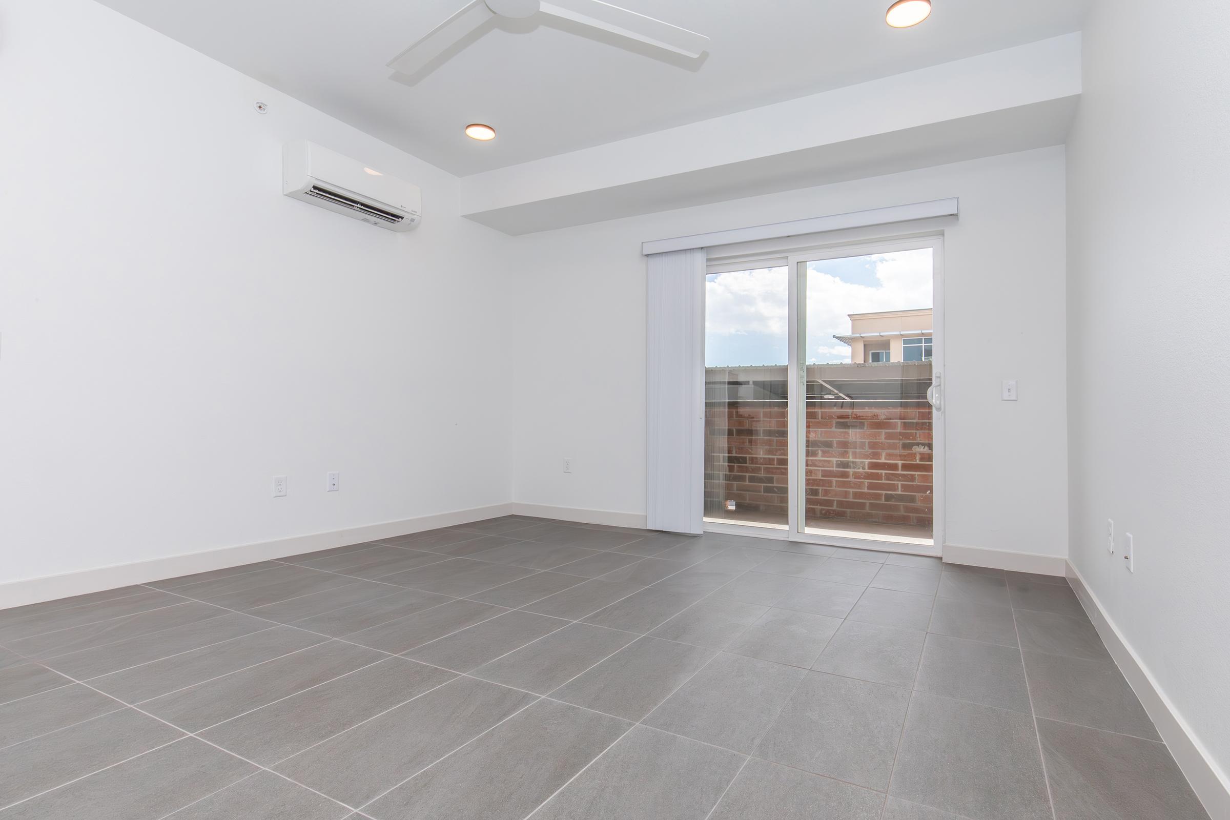 Spacious empty room with gray tile flooring, a ceiling fan, and large sliding glass doors that lead to a balcony. The walls are white, and there is a wall-mounted air conditioning unit. Natural light filters in through the doors, highlighting the room's clean, modern design.