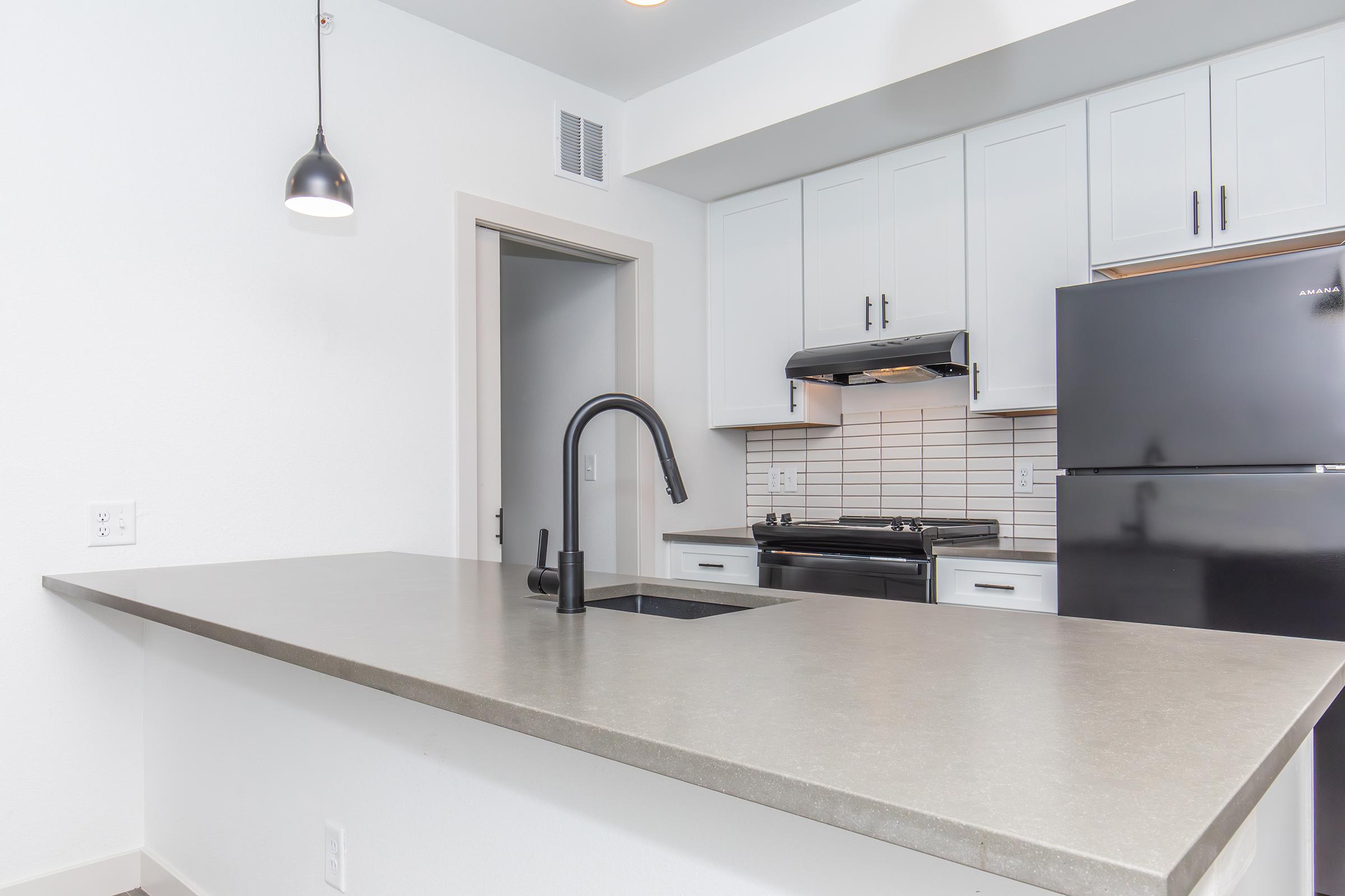 Modern kitchen featuring a concrete countertop, white cabinetry, and black appliances, including a refrigerator and stove. A sleek black faucet is installed in the sink, and there is a pendant light hanging above the counter. The walls are painted white, creating a bright and airy atmosphere.