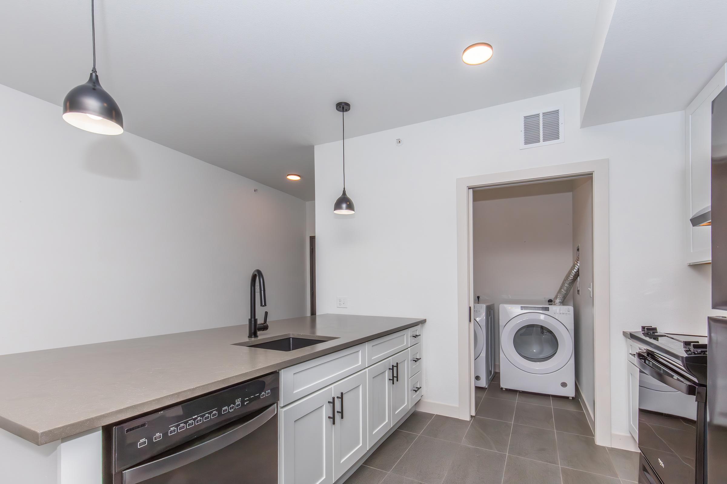 Modern kitchen featuring a gray countertop, stainless steel appliances, and white cabinetry. A sink with a black faucet is centrally located. Pendant lights hang from the ceiling. In the background, there is a laundry area with a washer and dryer. The space has a clean, contemporary design and neutral color palette.