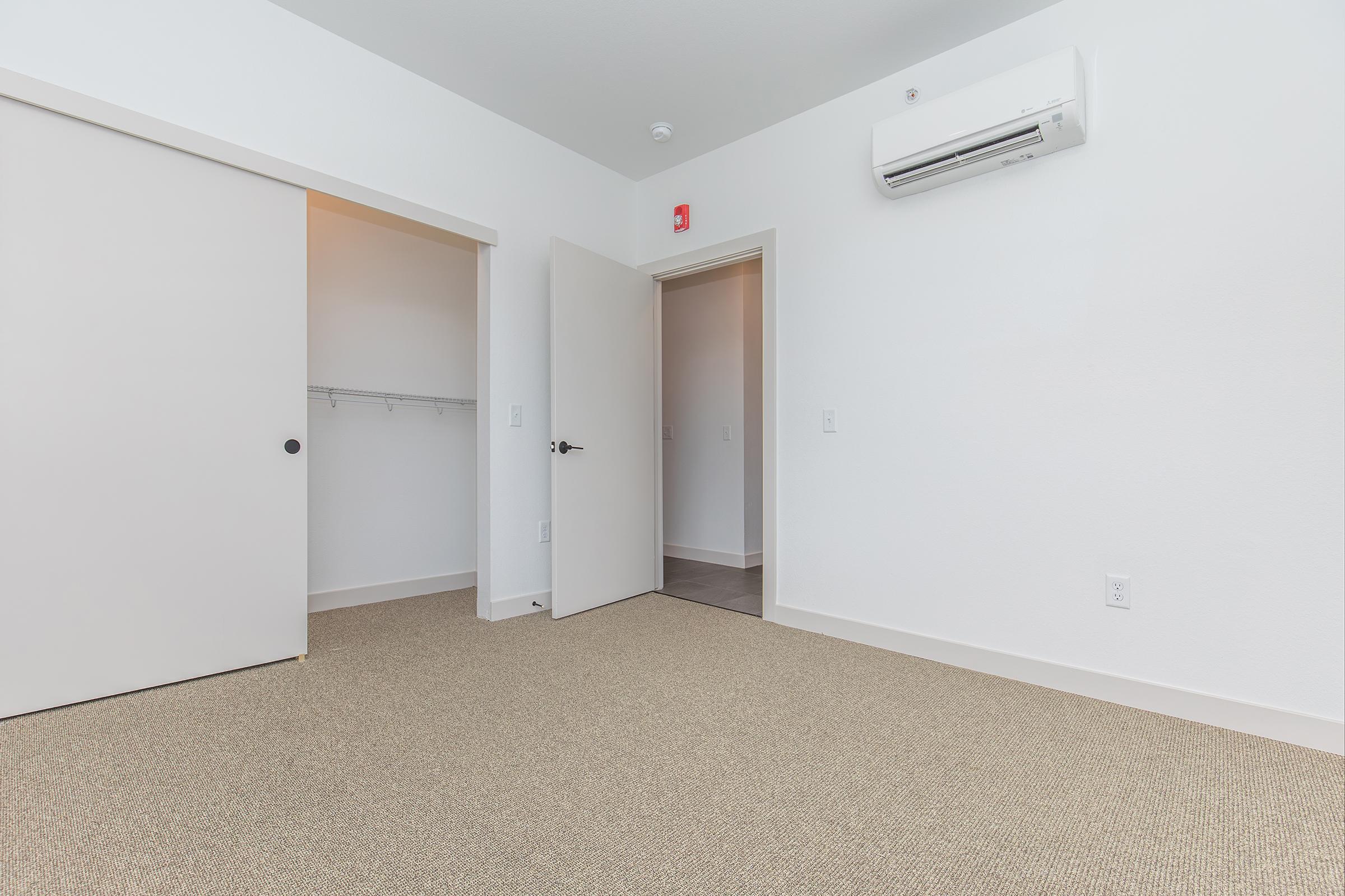 Empty room featuring light beige carpet, white walls, and a closet with sliding doors. A doorway leads to an adjacent space, and a wall-mounted air conditioner is visible. The overall atmosphere is clean and modern.