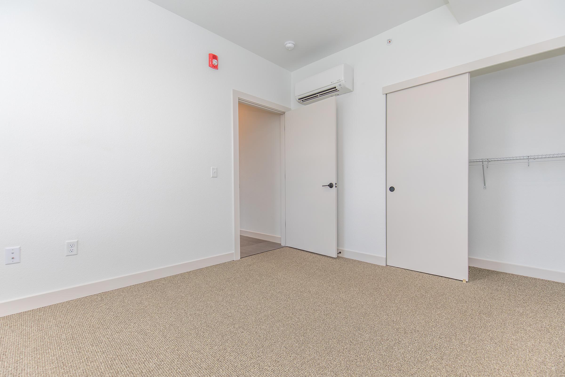 An empty room with beige carpet flooring and white walls. There are two doors: one leading to a closet with a hanging rod and the other is an entry door. An air conditioning unit is mounted on the wall. The space is well-lit with a simple, modern design.