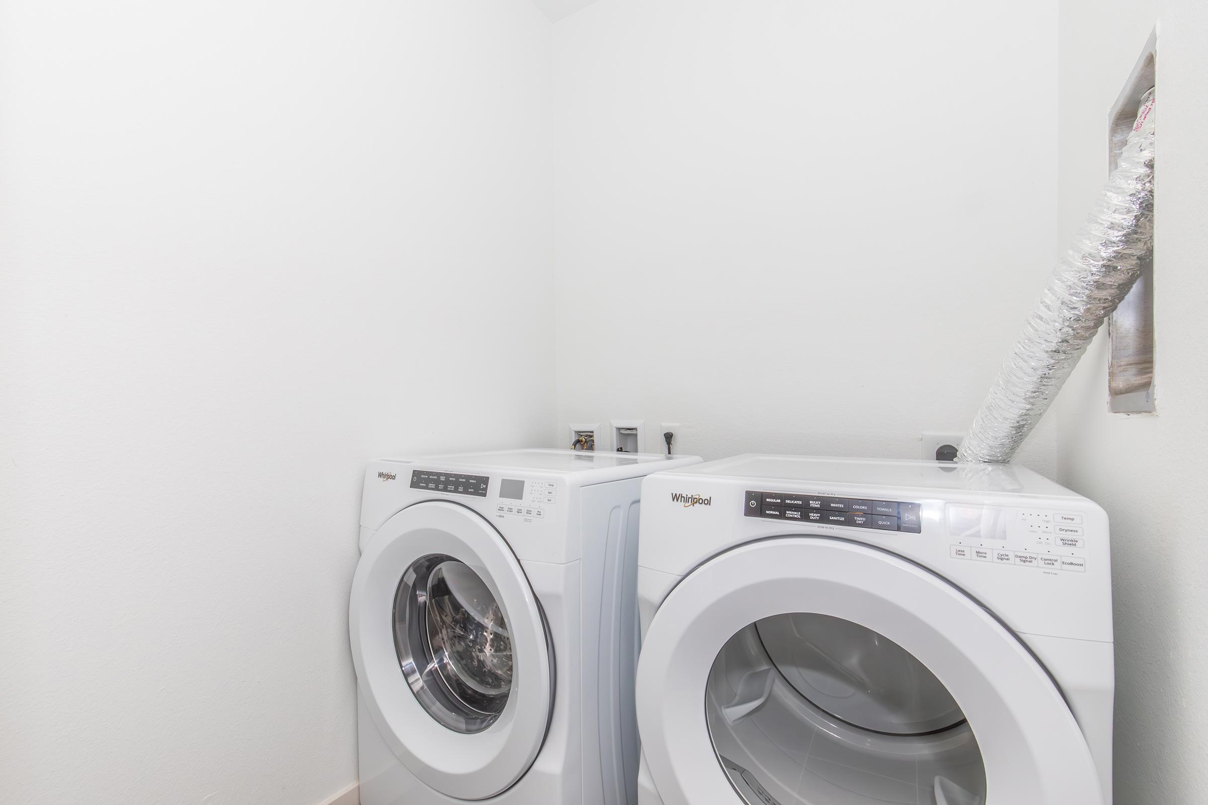 A bright laundry room featuring a set of white front-loading washing machines and a dryer side by side against a plain white wall. The room appears clean and well-maintained, with a vent and plumbing visible above the appliances.