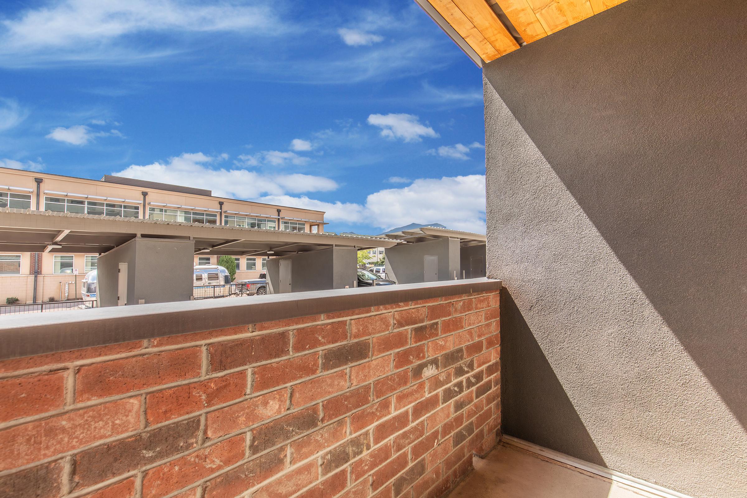 View from a balcony featuring a brick wall and a clear blue sky. In the background, there are several buildings with covered parking spaces, creating a sunny and open atmosphere. The scene captures a blend of architectural elements and natural lighting.