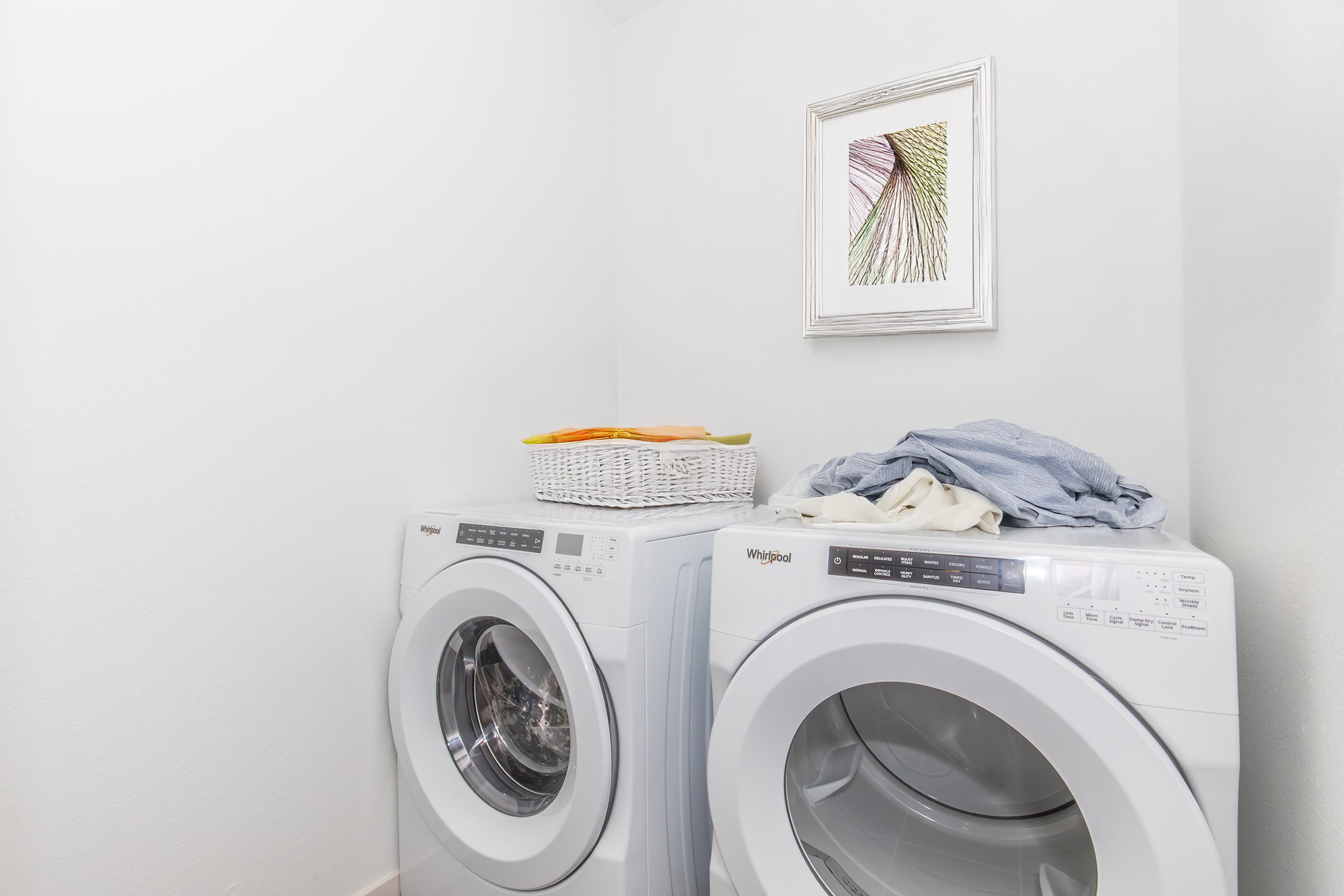 A modern laundry room featuring a pair of white washing machines stacked side by side. On top of the machines, there is a basket of laundry and a pile of clothes. The wall behind them is painted white, and there is a framed artwork above, depicting a leaf design, adding a touch of decor to the space.
