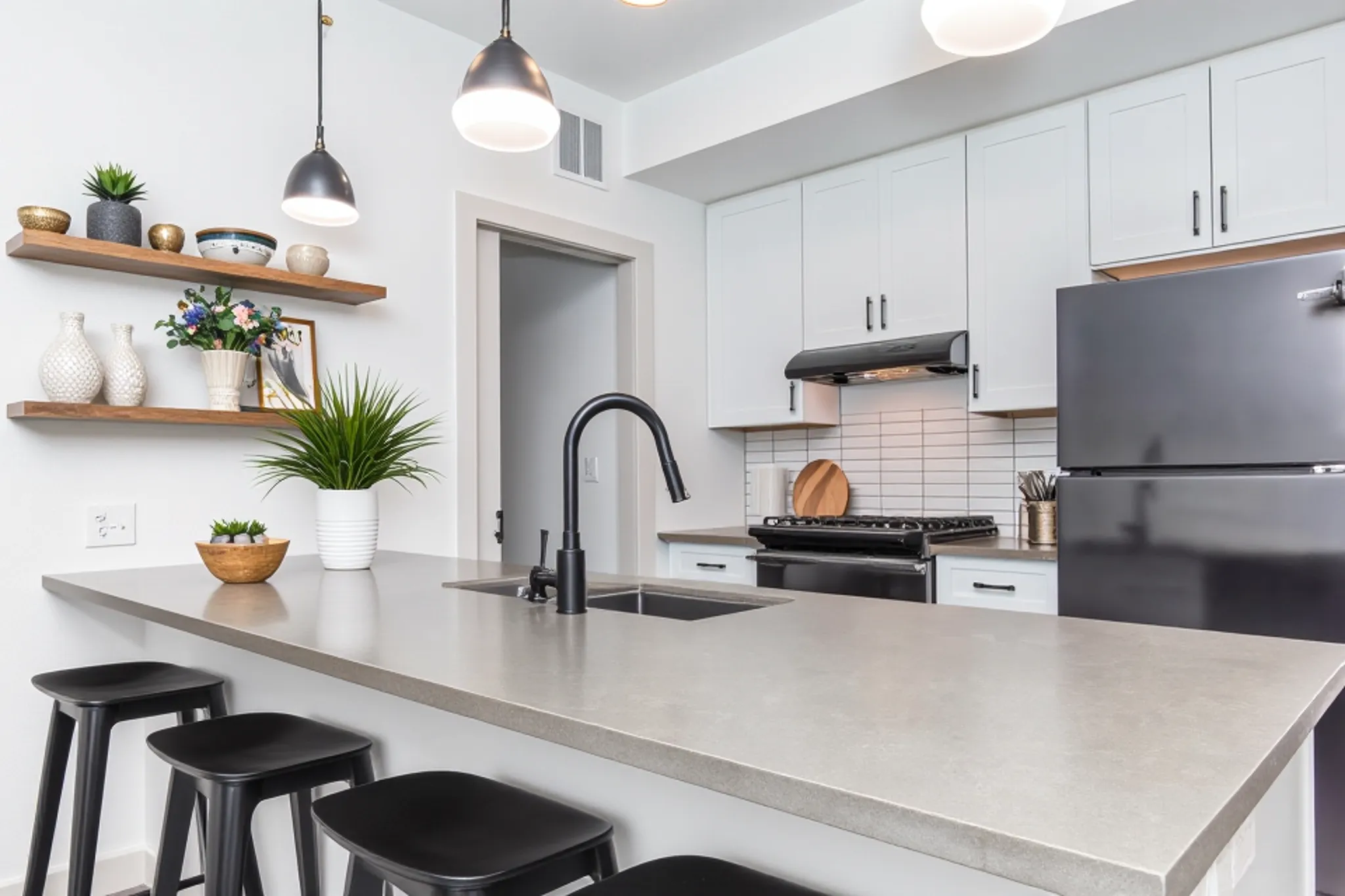 Modern kitchen featuring a minimalist design with light gray cabinets, black appliances, and a concrete countertop. A black faucet is installed over a sink with four black bar stools nearby. Shelves hold decorative vases and a plant, adding a touch of greenery to the space. Soft lighting from pendant lamps enhances the ambiance.