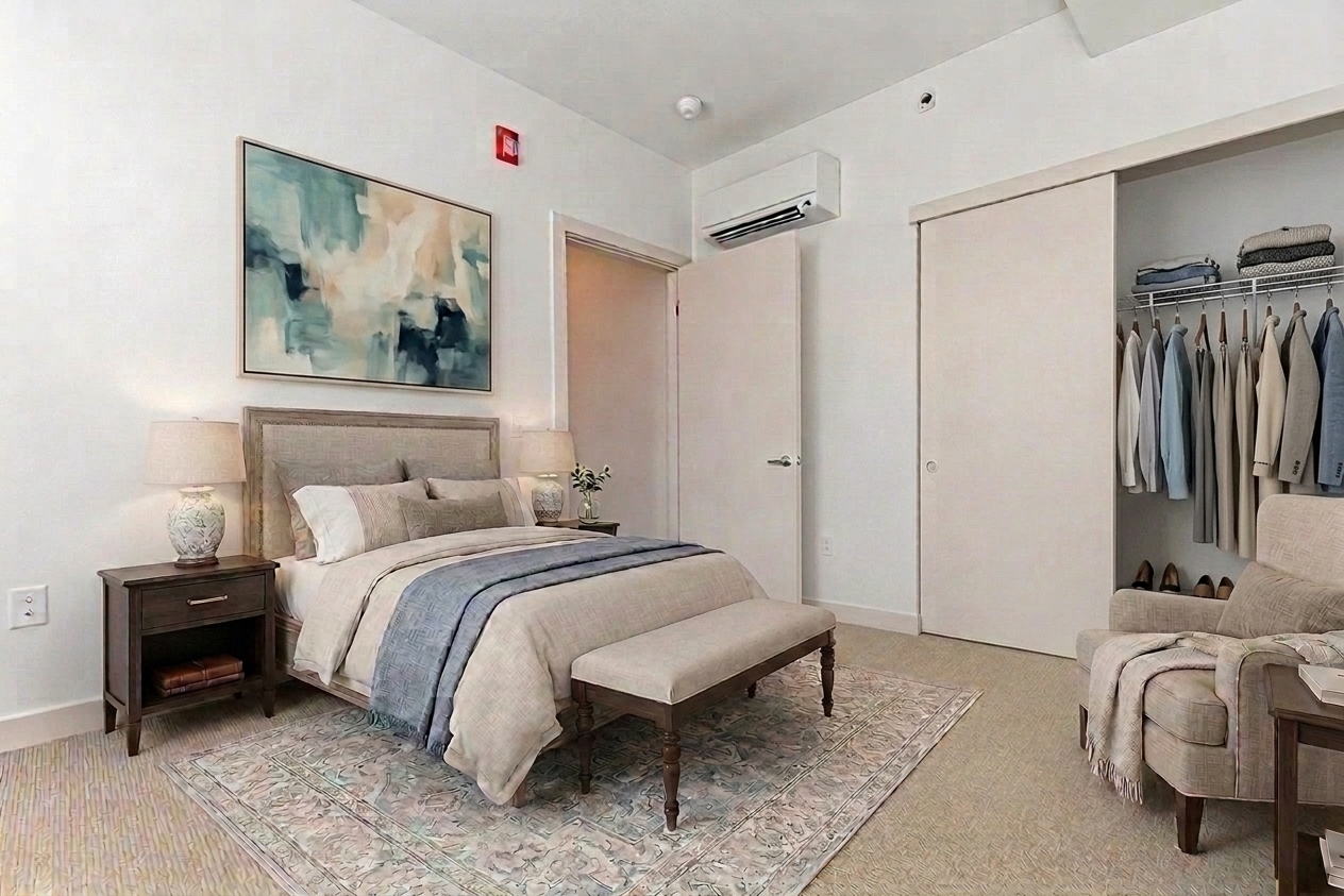 A cozy bedroom featuring a comfortable bed with a decorative blanket, a bedside table with a lamp, and an armchair. The walls are painted in neutral tones, and there is a piece of abstract artwork above the bed. A closet with hanging clothes is visible, along with a small rug adding warmth to the space.