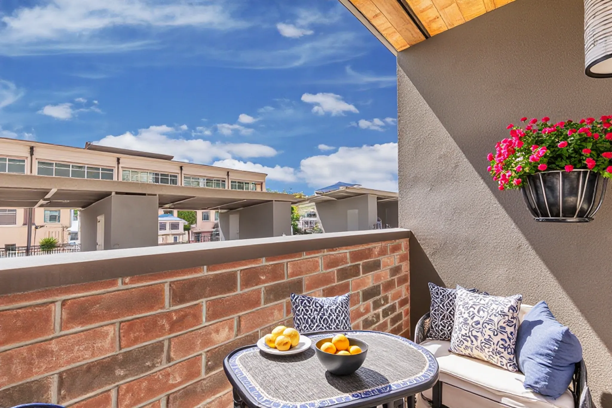 A cozy balcony with a small round table set for two, featuring a bowl of oranges and a decorative pillow on each chair. A flower pot hangs from the wall, filled with vibrant pink flowers, while a blue sky with fluffy clouds is visible in the background, along with a row of modern buildings.