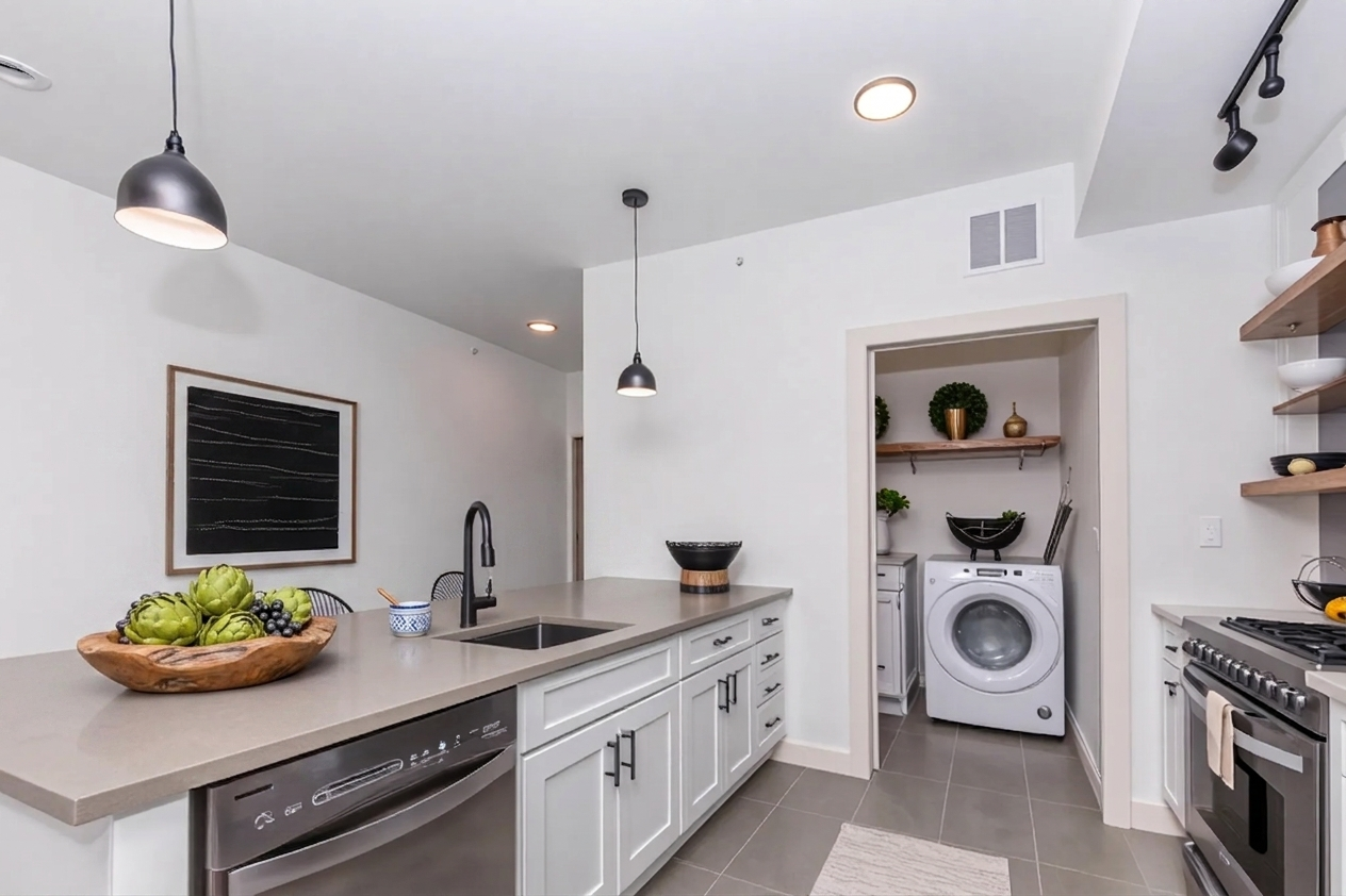 A modern kitchen featuring gray cabinets, a large island with a fruit bowl, stainless steel appliances, and an open laundry area with a washer. Pendant lighting hangs above the island, and decorative plants are displayed on shelves. The overall aesthetic is clean and contemporary.
