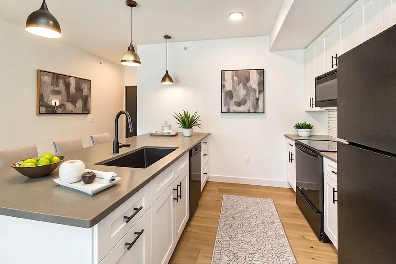 Modern kitchen featuring a central island with a dark countertop and sink. White cabinets line the walls, complemented by a black refrigerator and oven. Pendant lights hang above the island, and artwork is displayed on the walls. A bowl of green apples and a small plant add decorative touches to the space.