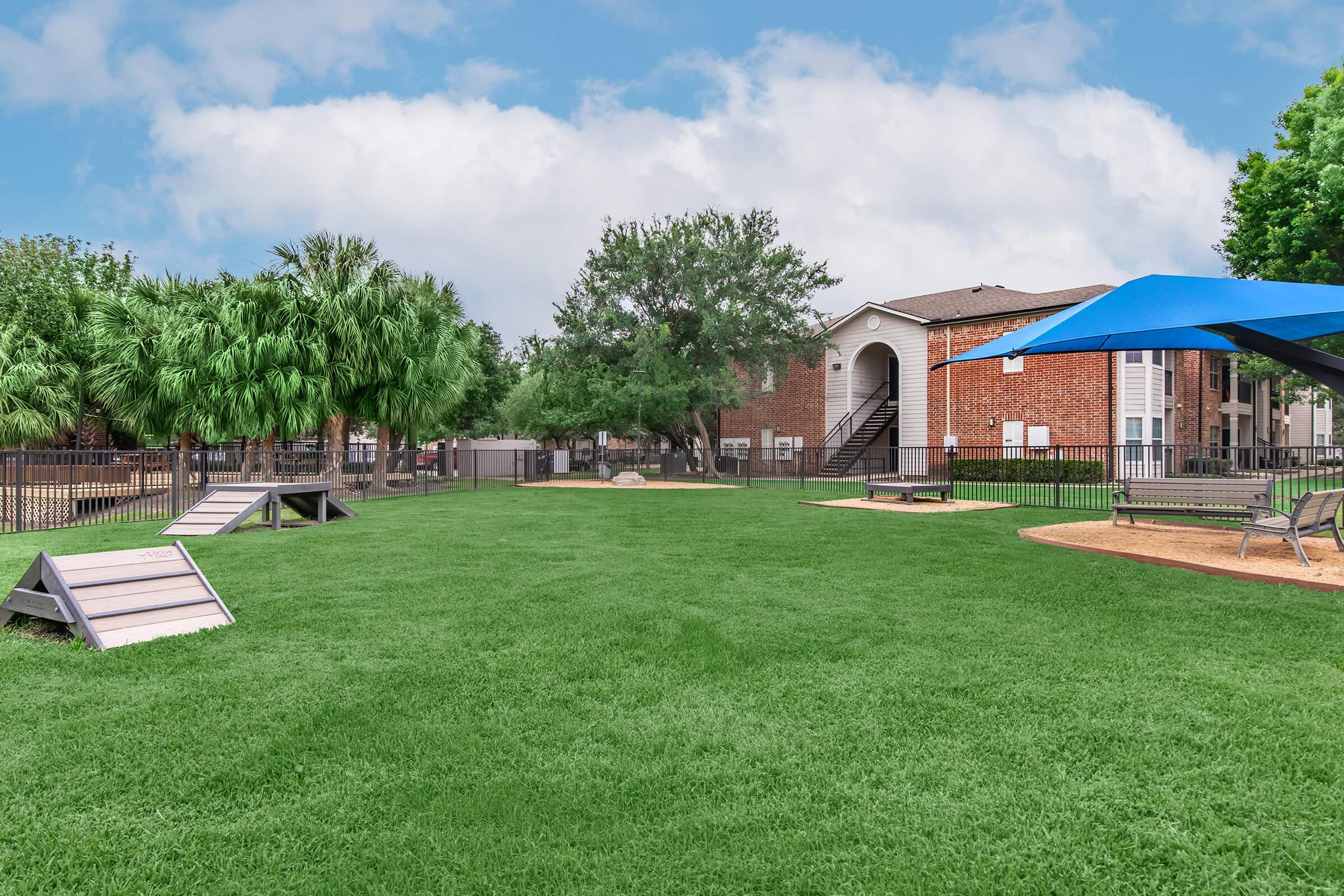 A spacious green lawn area featuring playground equipment and shaded benches. Surrounding trees provide a natural ambiance, and a nearby apartment building is visible in the background, along with a blue shade structure. The scene conveys a relaxed outdoor space ideal for recreation or leisure.