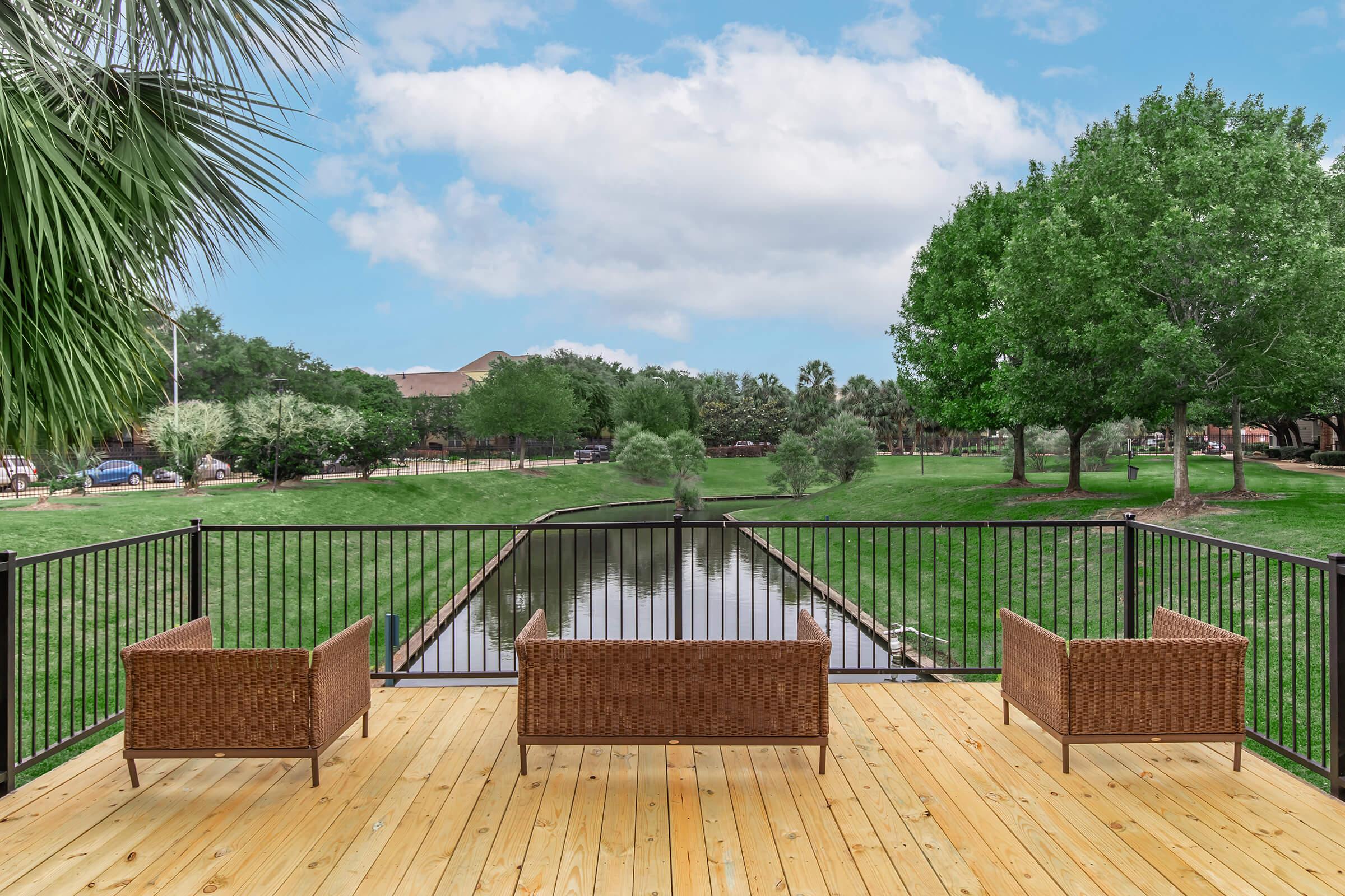 A wooden deck with three wicker chairs overlooks a peaceful scene featuring a calm waterway and lush greenery. Trees and shrubs line the area, offering a serene outdoor setting under a partly cloudy sky.