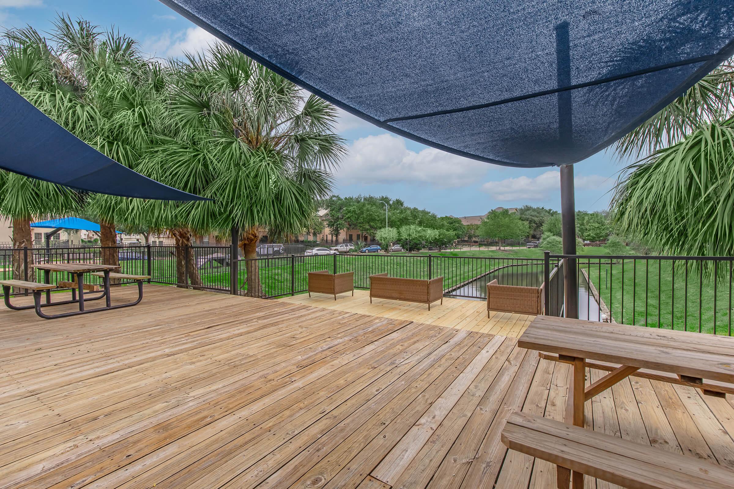 Outdoor patio area with wooden flooring, featuring picnic tables and shaded seating under large blue canopies. Lush palm trees are in the background, with a view of a green lawn and water. The sky is partly cloudy, creating a pleasant, relaxing atmosphere.