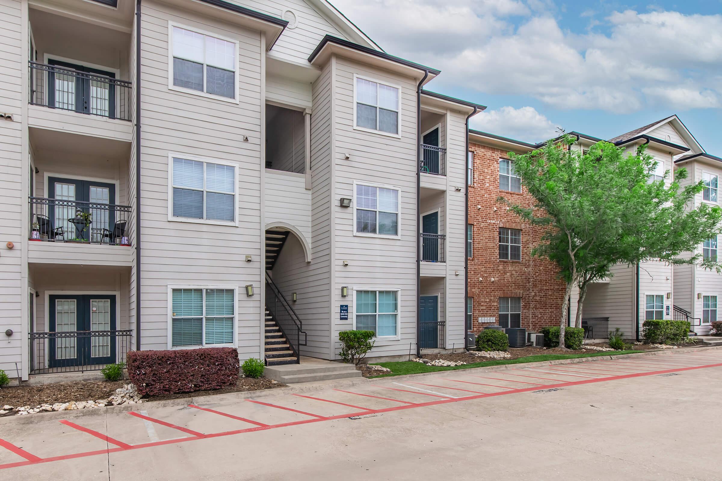 Exterior view of a multi-story apartment complex featuring two distinct buildings, one with light-colored siding and balconies, and another with brick facade. The scene includes a landscaped area, parking spaces, and trees, with a cloudy sky in the background.
