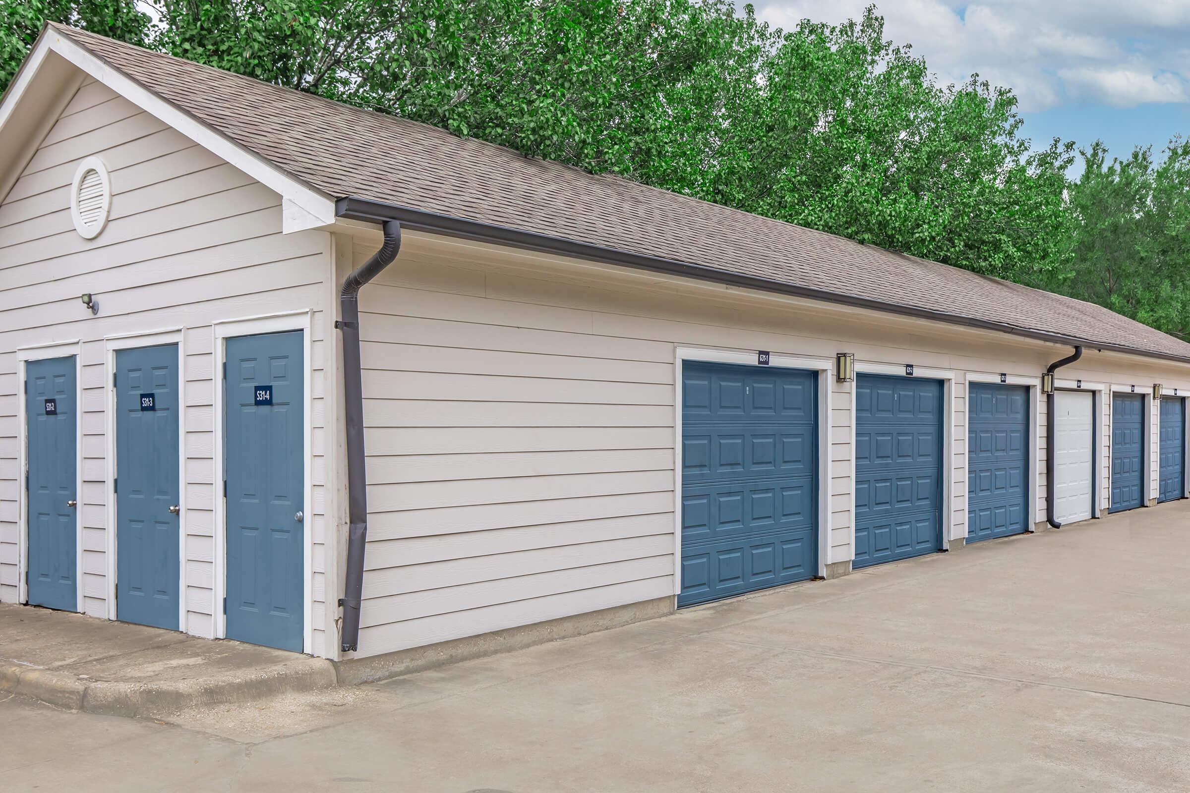 A row of storage units with blue doors and light-colored siding, situated beside a paved path. Lush green trees are visible in the background, under a partly cloudy sky.