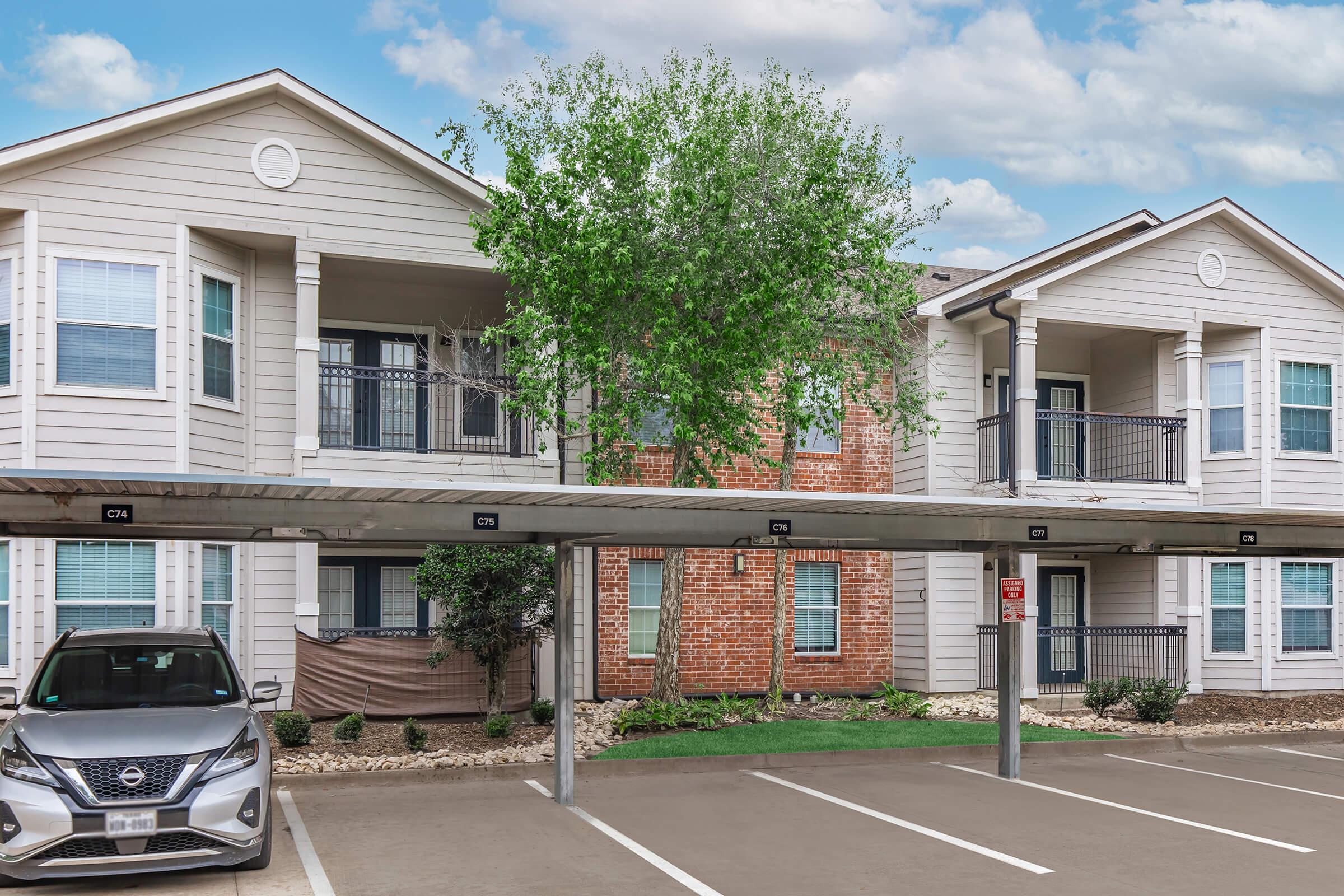 A residential apartment complex featuring two buildings with balconies. In the foreground, there is a car parked under a covered carport. The area includes landscaped greenery, and the sky is partly cloudy.