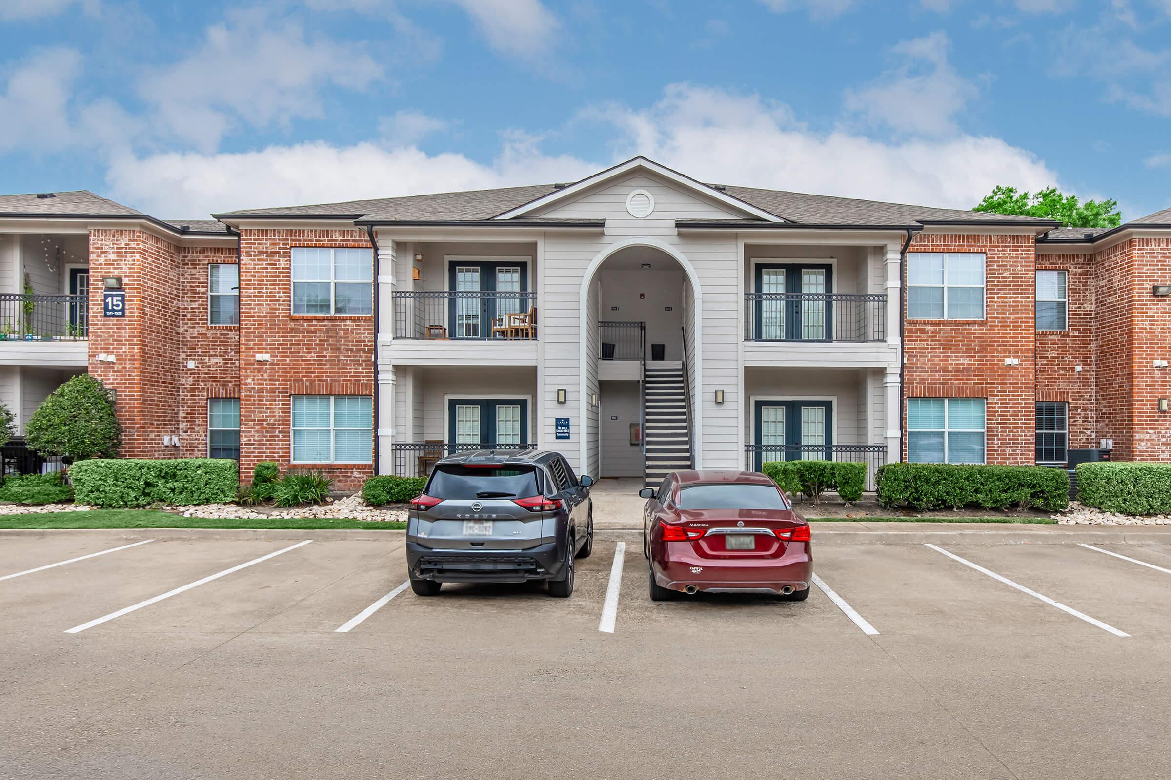 A two-story brick apartment building with white trim, featuring balconies on the upper level. Two parked cars, a dark SUV and a red sedan, are in the foreground. There are neatly trimmed bushes and a paved parking lot in front of the building, under a partly cloudy sky.