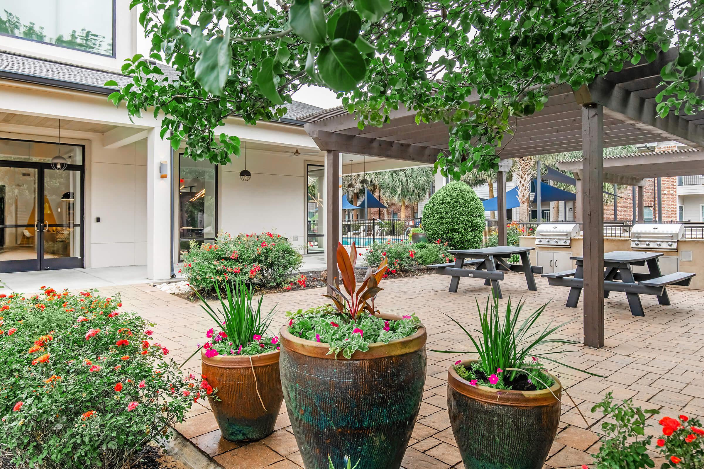 A landscaped outdoor area featuring vibrant flower pots with colorful plants, a shaded seating area with picnic tables, and a barbecue grill station. In the background, there are palm trees and a swimming pool visible, creating a relaxing atmosphere.