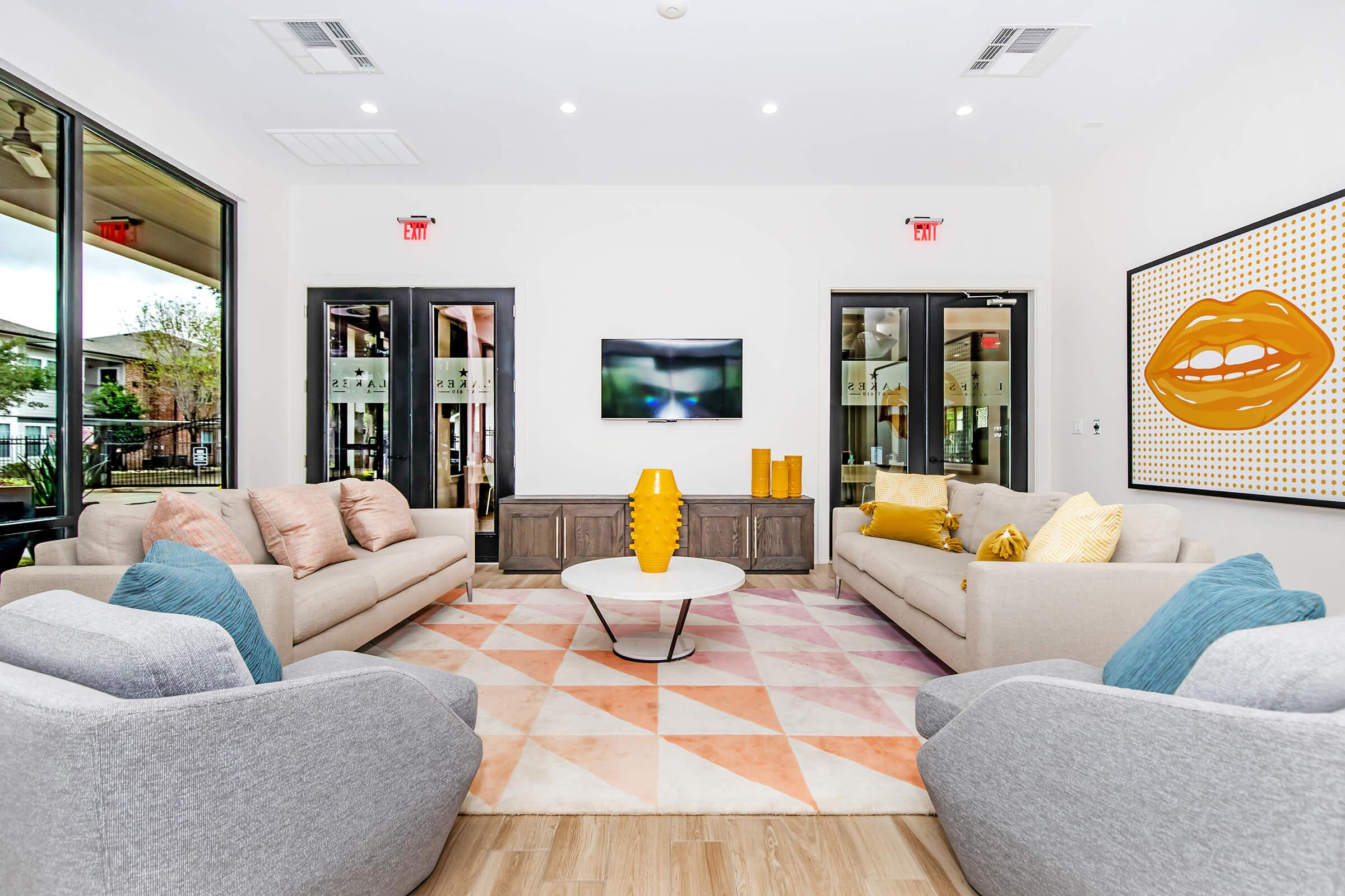 A modern living room featuring two gray armchairs and two light-colored sofas adorned with colorful throw pillows. A round coffee table is centered on a geometric-patterned rug. A flat-screen TV mounted on the wall displays a blurred image, and large windows provide natural light, complementing the contemporary decor.