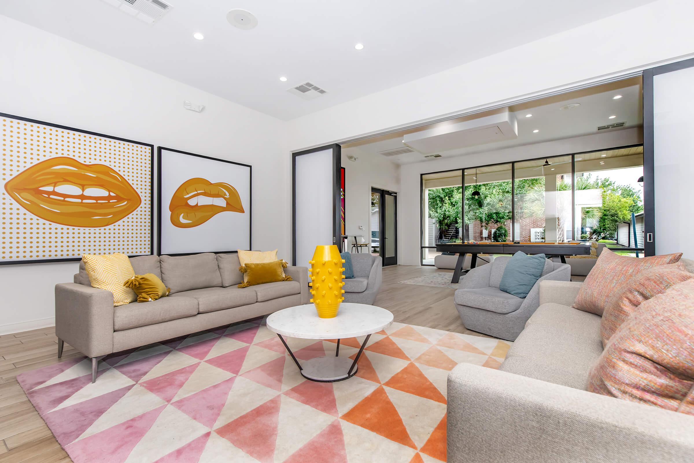 A modern living room featuring two stylish sofas, a round coffee table, and a colorful geometric rug. The walls are adorned with bold art pieces, and large windows allow natural light to fill the space, offering a view of greenery outside. Accents include vibrant cushions and a bright yellow vase on the table.