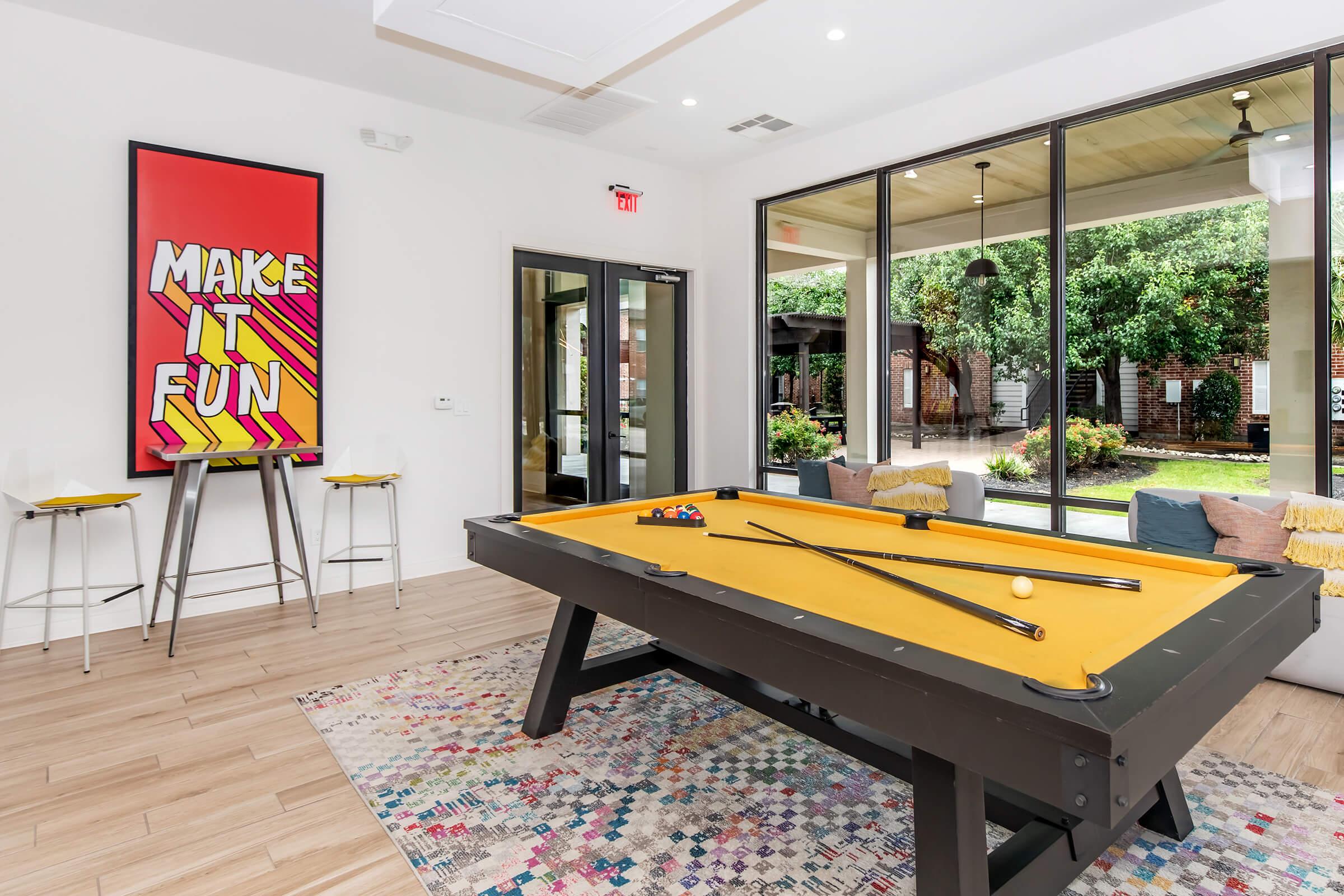 A vibrant game room featuring a bright yellow pool table with cues and balls. The walls are adorned with colorful artwork, including a bold "MAKE IT FUN" poster. Large windows provide natural light and a view of an outdoor area with seating, creating an inviting and playful atmosphere.