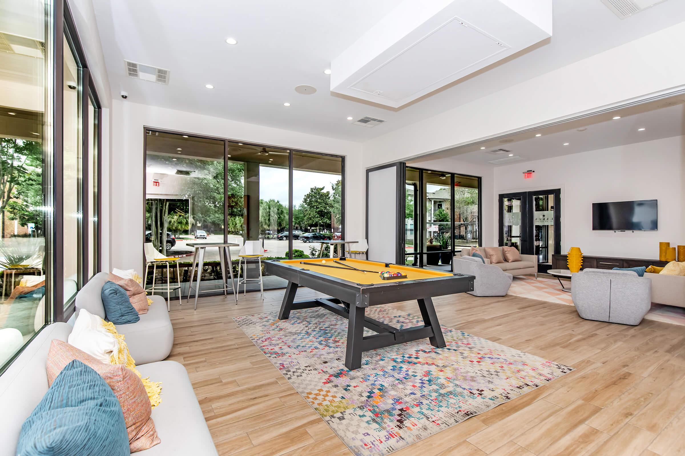 A modern communal lounge area featuring a billiard table at the center, surrounded by cozy seating on a colorful rug. Large windows provide natural light and outdoor views. A dining area is visible in the background with high chairs and tables, creating a welcoming atmosphere for relaxation and socializing.