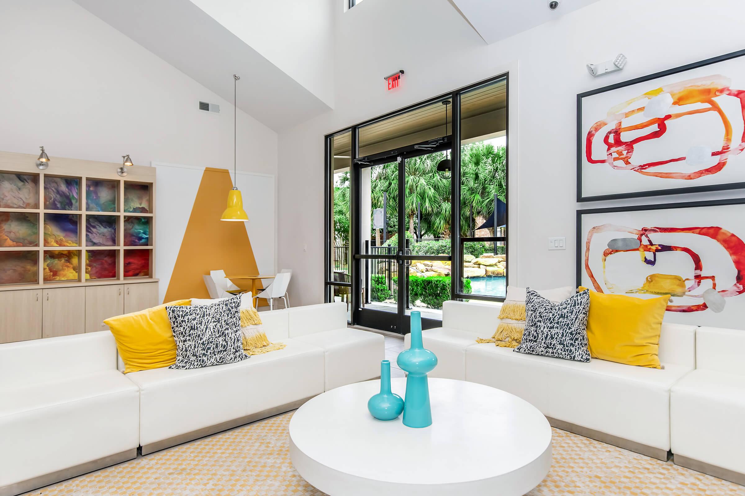 A modern living room featuring a white sectional sofa adorned with yellow and patterned cushions. A round white coffee table sits in the center, with turquoise decorative vases. Behind, large windows provide a view of lush greenery, and colorful artwork is displayed on the walls. Bright and airy atmosphere.