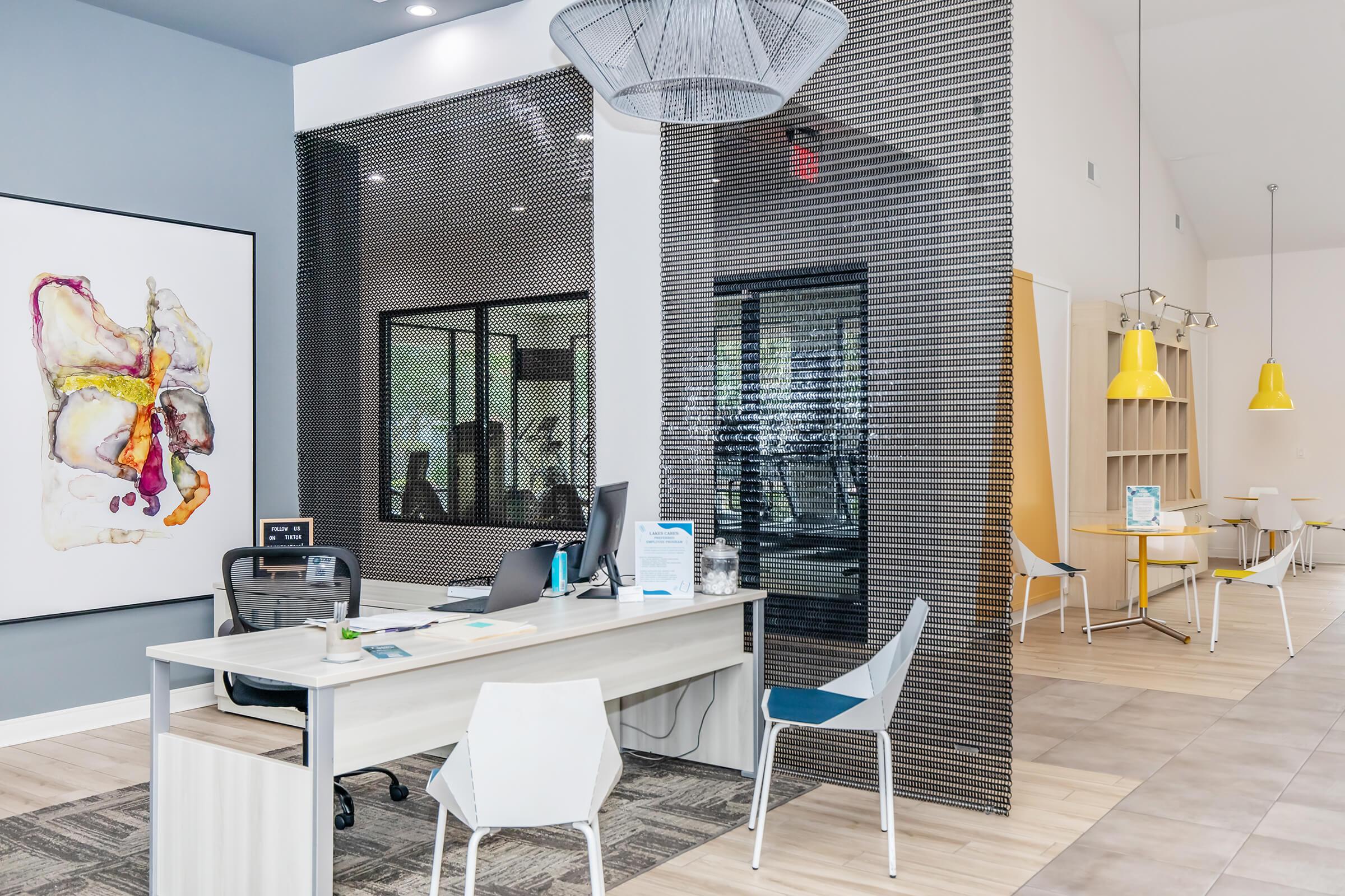 Modern office reception area featuring a sleek desk with a computer, stylish chairs, and artistic wall decor. The space is bright and inviting, with large windows, colorful pendant lights, and a contemporary design. A glass partition adds a touch of sophistication to the environment.