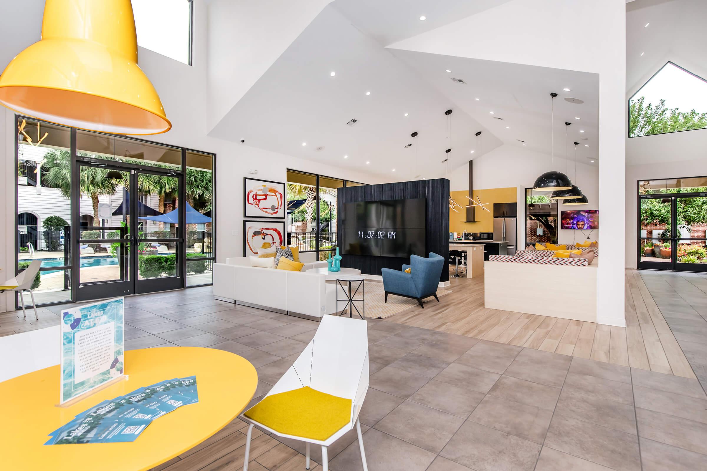 A modern, spacious lobby area featuring a large open layout with stylish furniture. Highlights include a bright yellow pendant light, a white and gray seating area, a large TV, and a communal dining space. Large glass doors lead to an outdoor pool area, creating a welcoming and vibrant atmosphere.