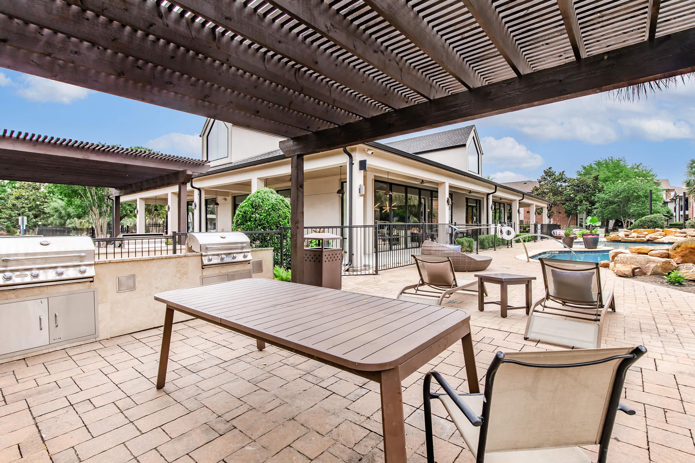 Outdoor patio area featuring a table and chairs, a built-in grill, and a large countertop. In the background, a modern building with large windows is visible, surrounded by well-maintained greenery. A swimming pool is partially seen, creating a relaxing atmosphere.