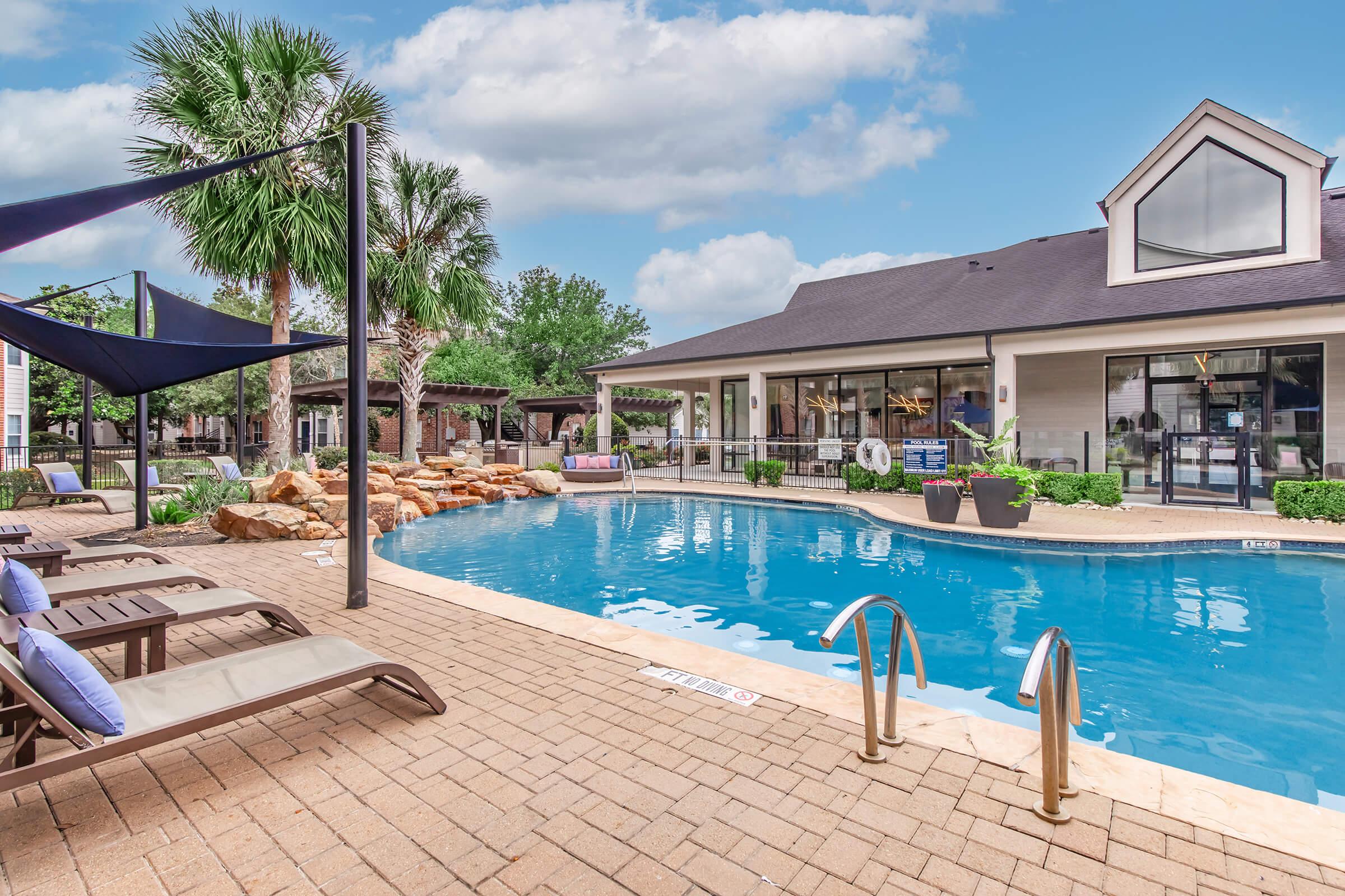 A tranquil outdoor swimming pool surrounded by lounge chairs, palm trees, and landscaped rocks. The backdrop features a modern building with large windows, offering a relaxing atmosphere. A few decorative cushions and shade structures enhance the inviting scene. Bright, clear skies contribute to the serene ambiance.