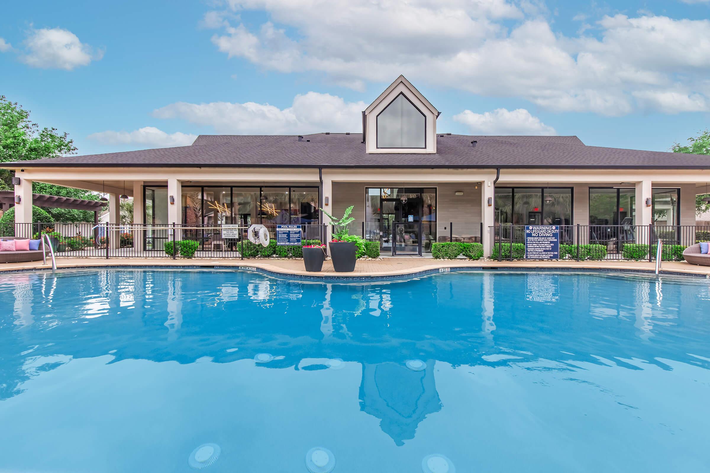 A well-maintained swimming pool in front of a modern building with large windows. The pool area features lounge chairs and greenery, creating a welcoming atmosphere. Clear blue skies and fluffy clouds enhance the scene, suggesting a sunny day.
