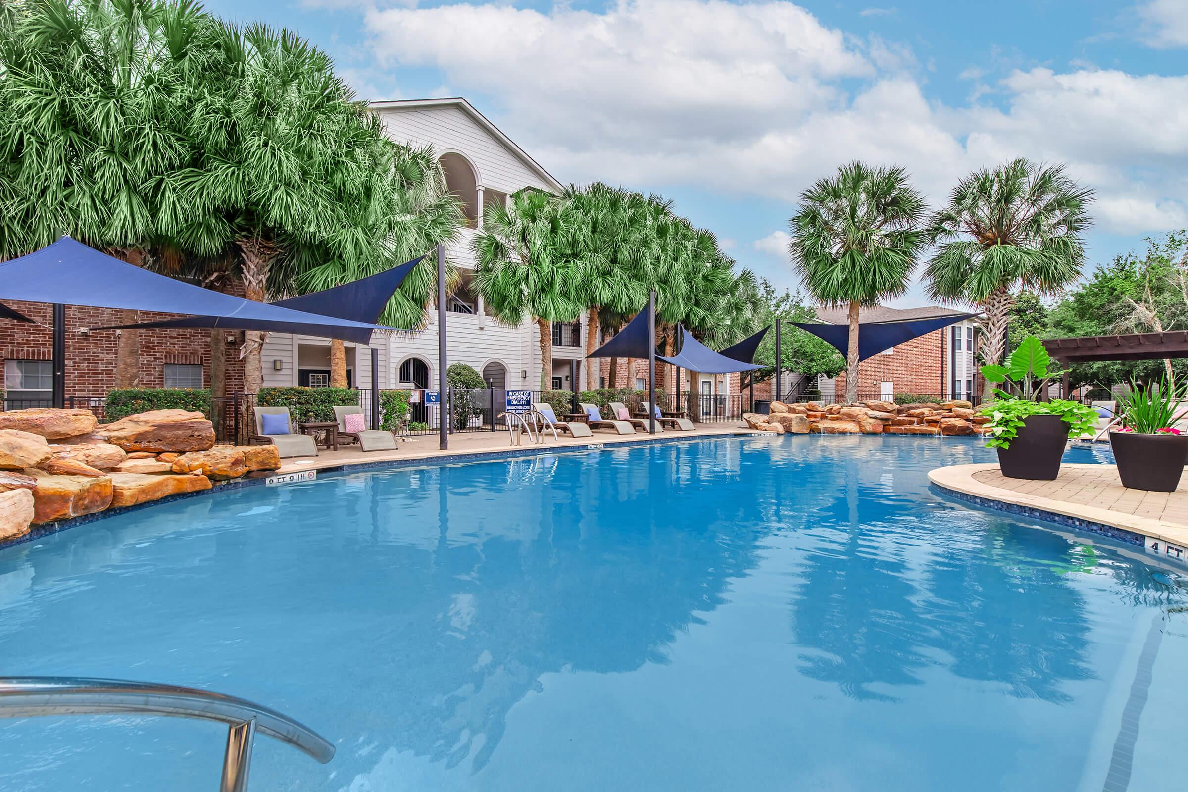 A serene outdoor pool area surrounded by lush palm trees and tropical plants. The pool features a clear blue water surface, lounge chairs, and shaded areas with large umbrellas. The backdrop includes a multi-level apartment building with a light exterior. The scene conveys a relaxing and inviting atmosphere.