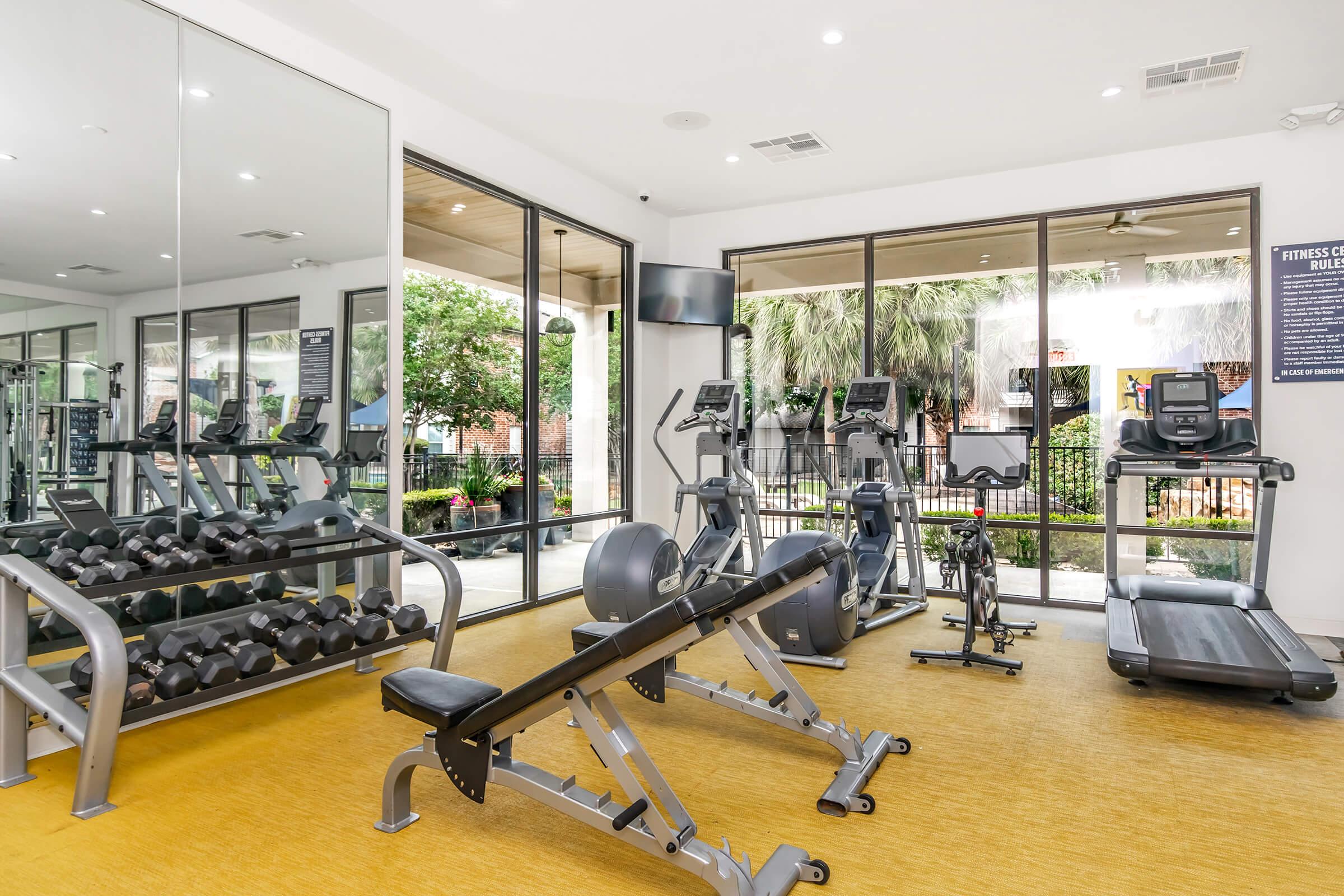A modern gym interior featuring various fitness equipment including dumbbells, a weight bench, a treadmill, and an exercise bike. Large windows allow natural light to fill the space, and potted plants are visible outside, creating a bright and inviting atmosphere for workouts.
