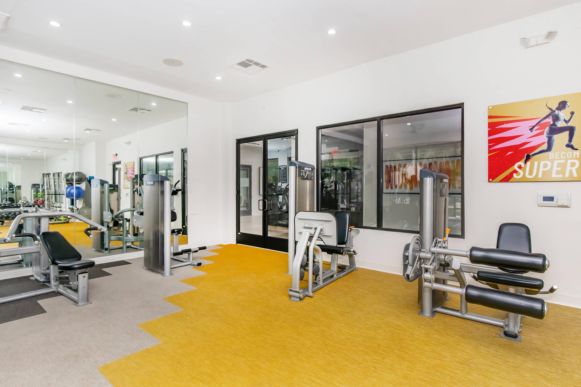 A modern gym interior featuring various workout machines, including strength training equipment. The bright space has large windows, reflective mirrors, and a vibrant yellow carpet. A motivational poster on the wall encourages fitness, and the atmosphere is well-lit and inviting for exercise.