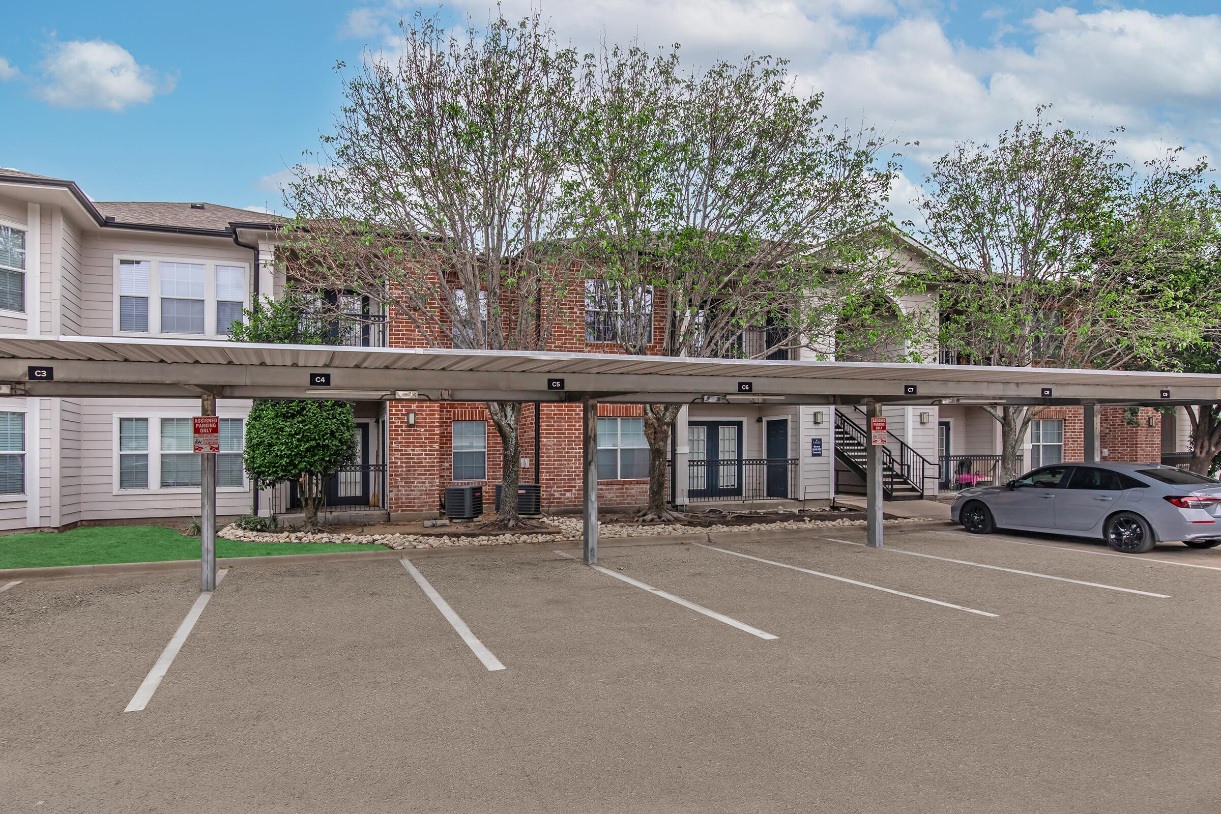 A parking area with several covered spaces in front of a two-story residential building. The building features brick and siding exteriors with small trees and greenery. Parked cars include a gray sedan. The environment appears well-maintained and inviting.