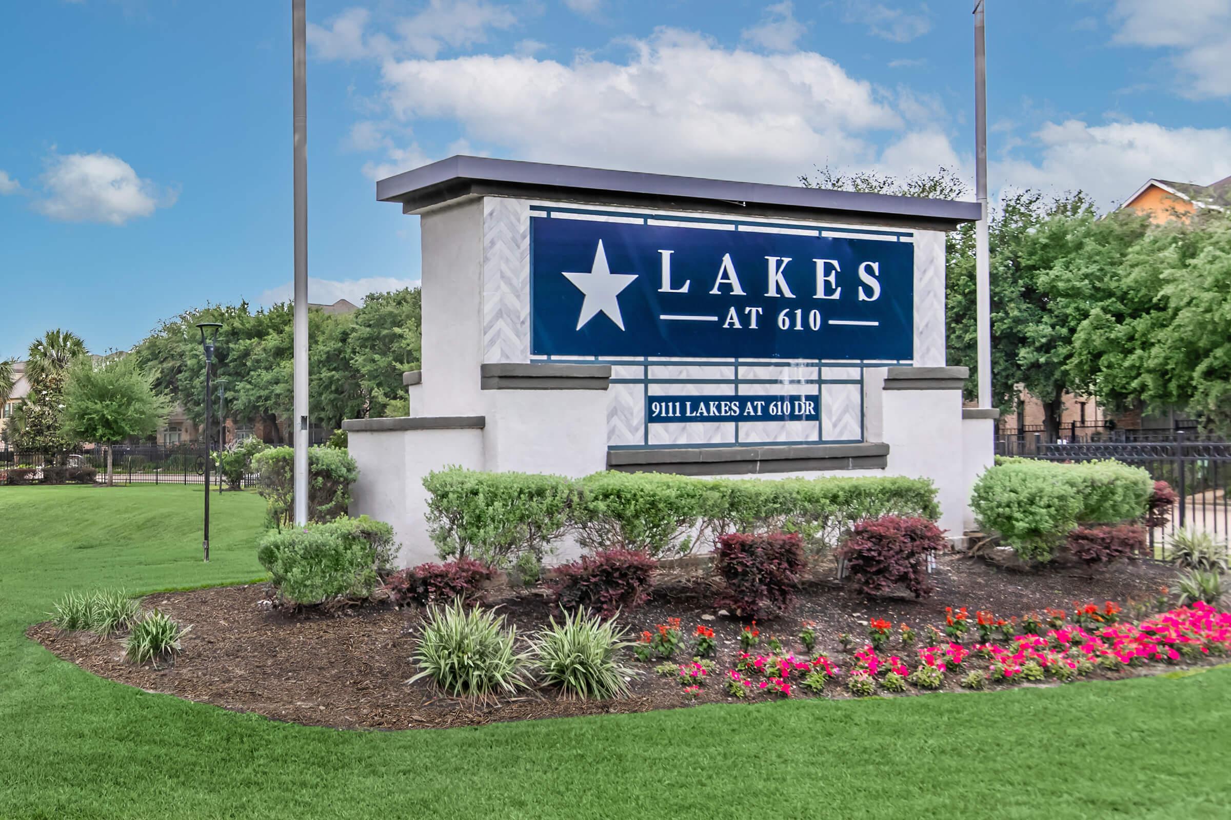 Sign for "Lakes at 610" featuring a blue background with a white star. The sign is surrounded by well-maintained landscaping, including green grass, shrubs, and colorful flowers. The sky is clear with some clouds, creating a bright and inviting atmosphere.
